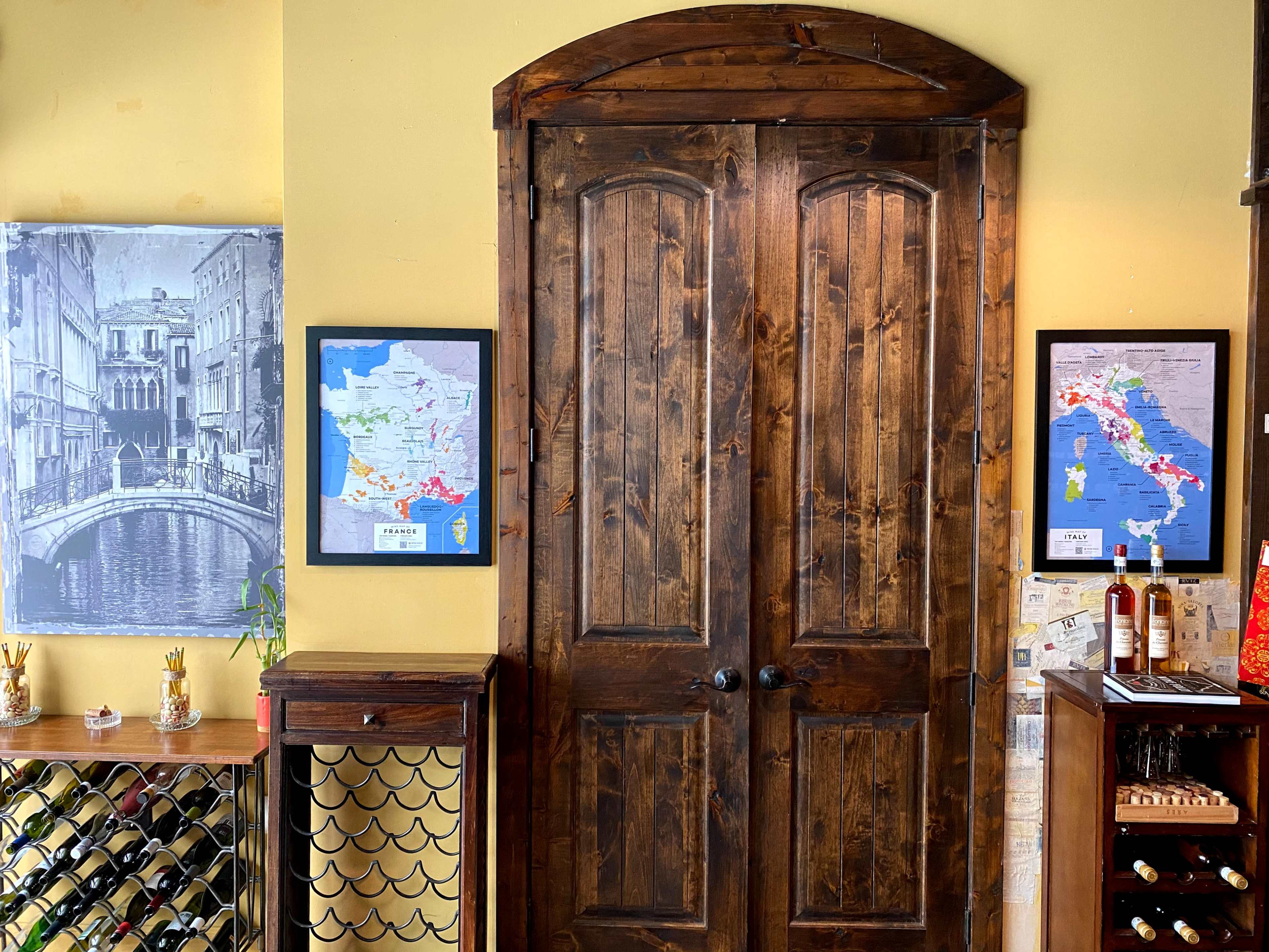 The scene features a rustic wooden door flanked by framed maps and a wine rack, set against a warm, yellow wall.