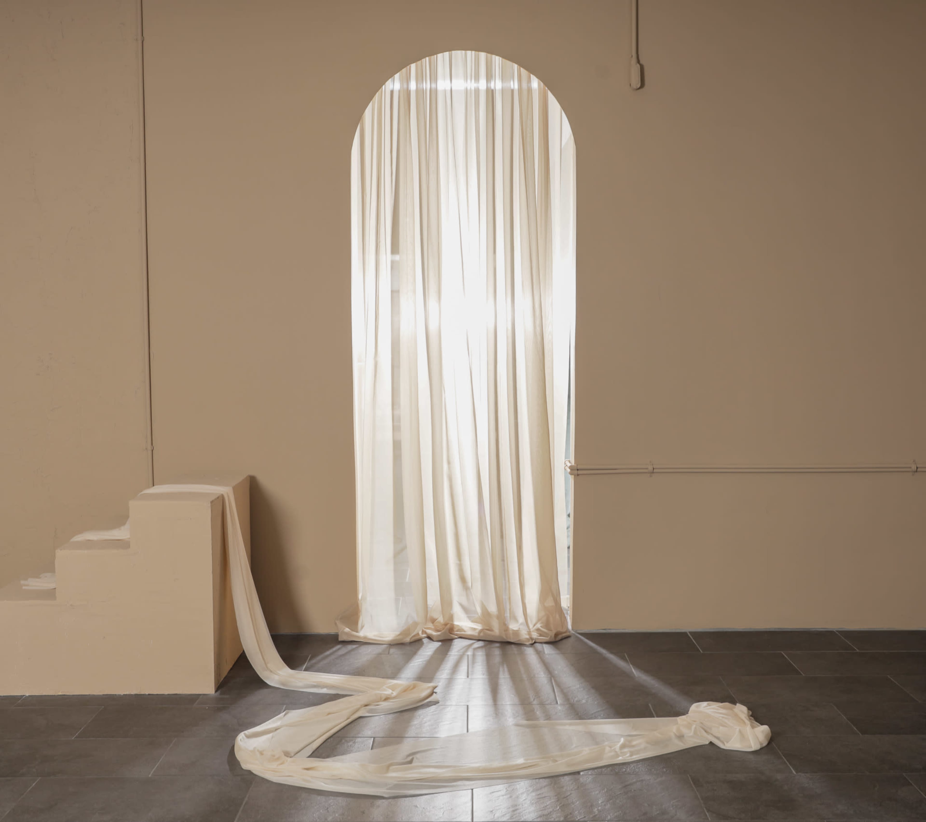 A sheer curtain drapes over an arched doorway, with soft fabric cascading along the floor beside a small staircase in a minimalist room.