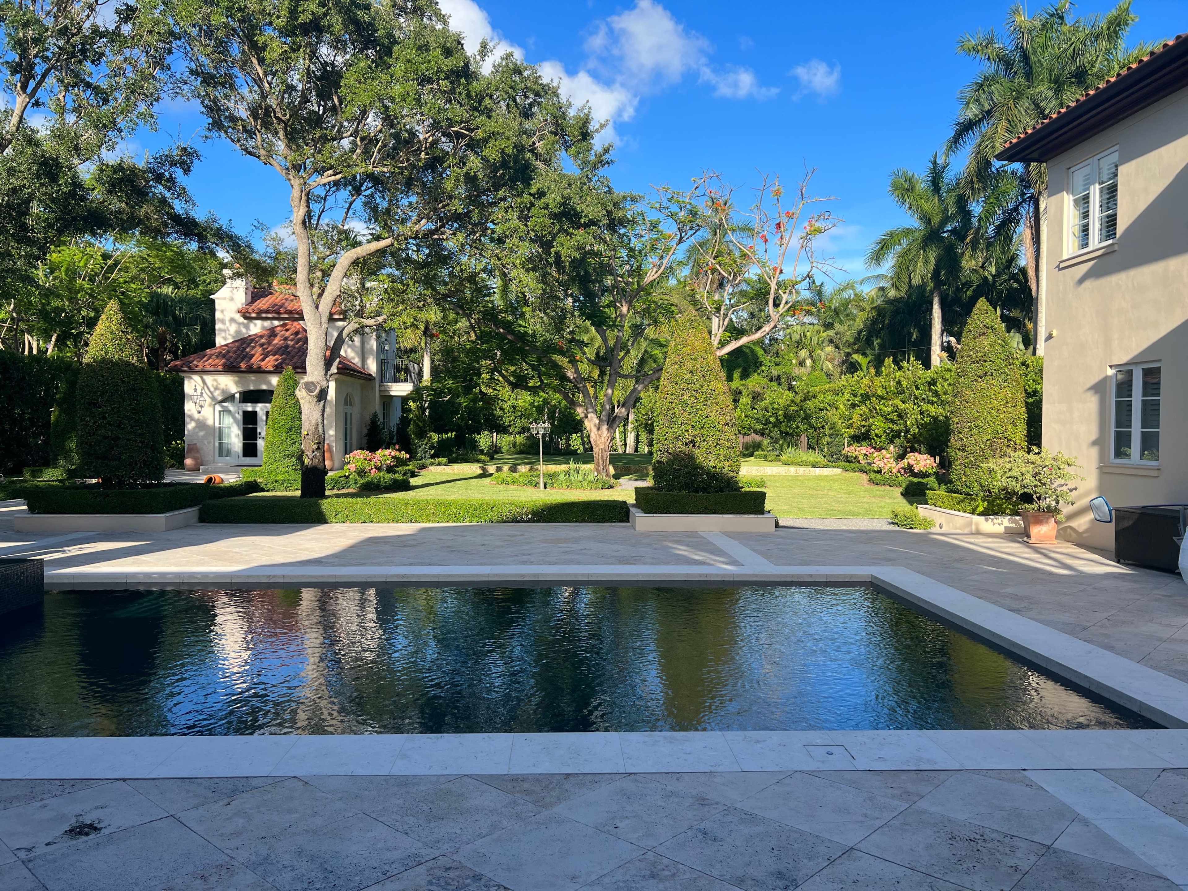 A landscaped backyard featuring a stone pool surrounded by lush greenery and trees, with a house visible on the right.