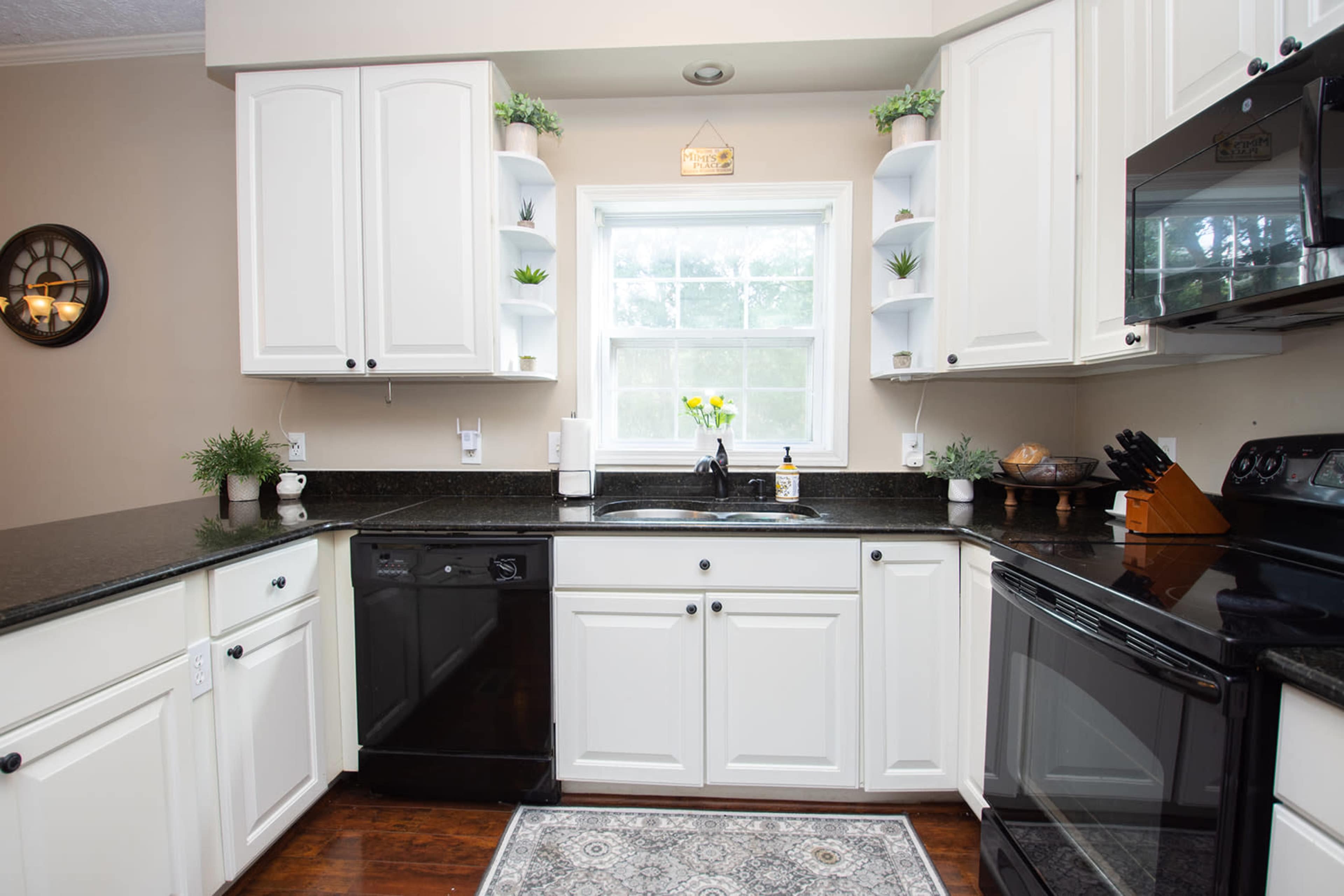 A modern kitchen with white cabinets, a dark countertop, and a window above a sink surrounded by plants and small decorative items.