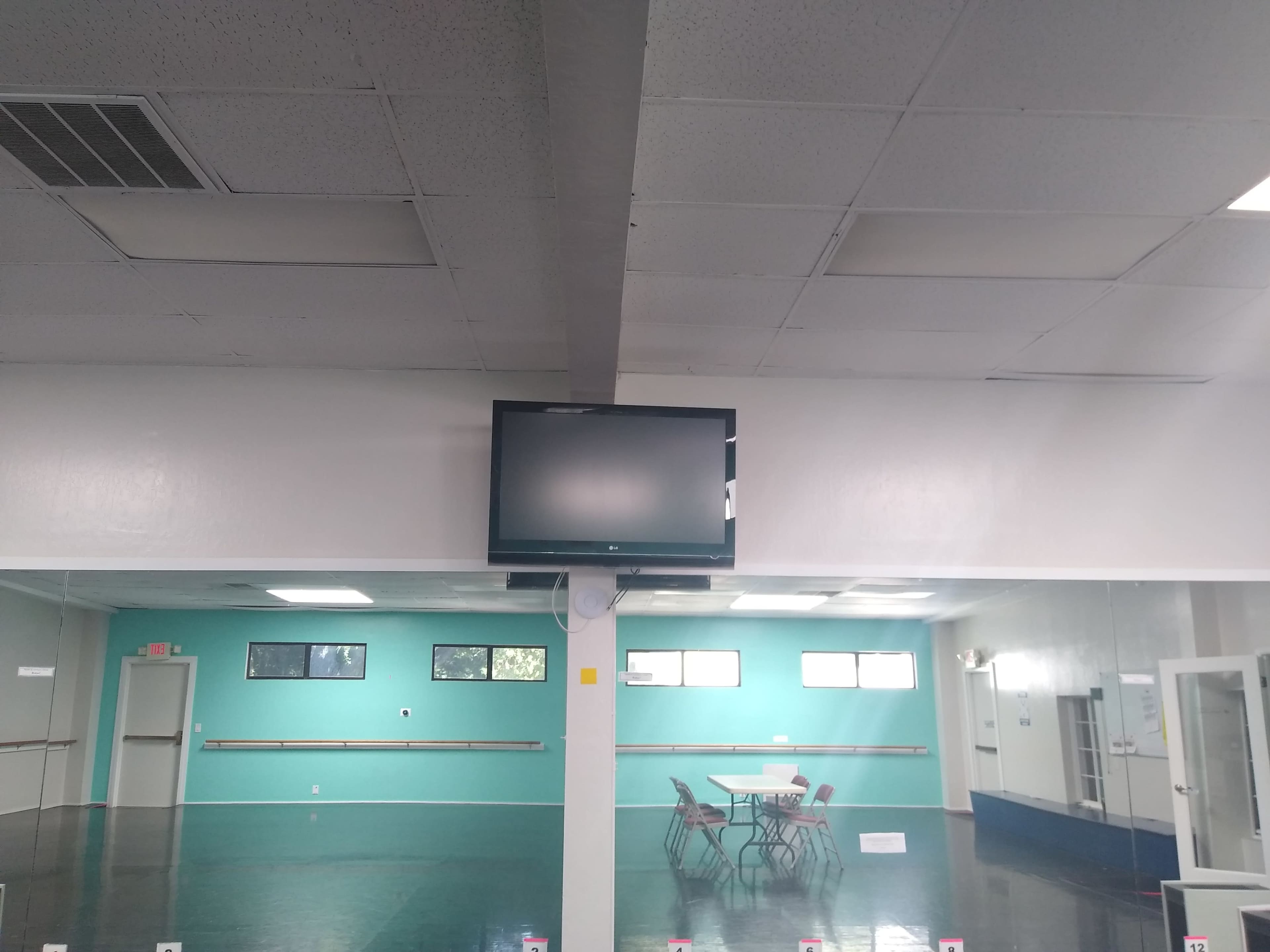 The image shows a spacious room with a dark floor, light green walls, a television mounted on the wall, and a table with chairs set up in the center.