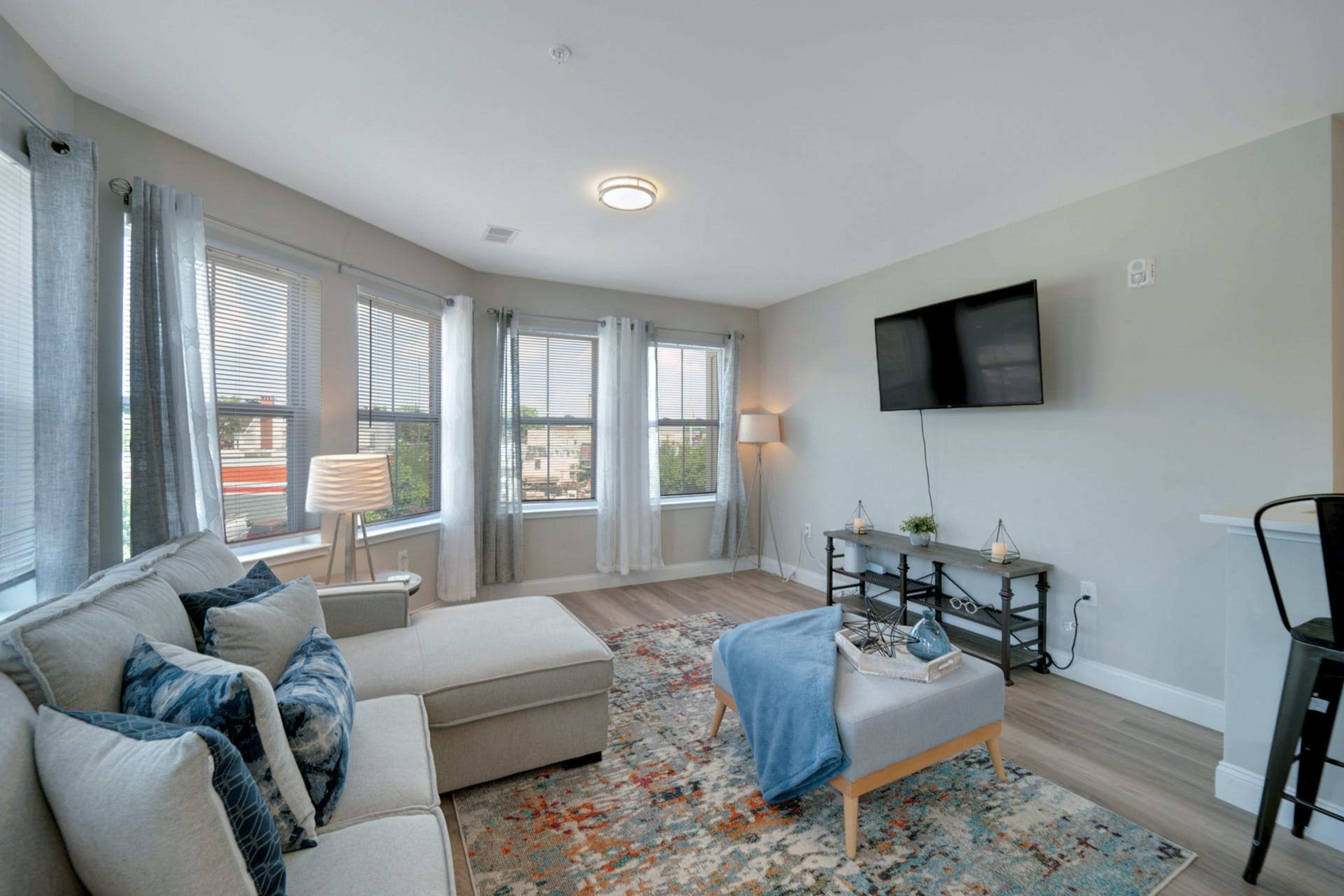 A modern living room features a beige sofa, a flat-screen TV mounted on the wall, and large windows allowing natural light to enter.