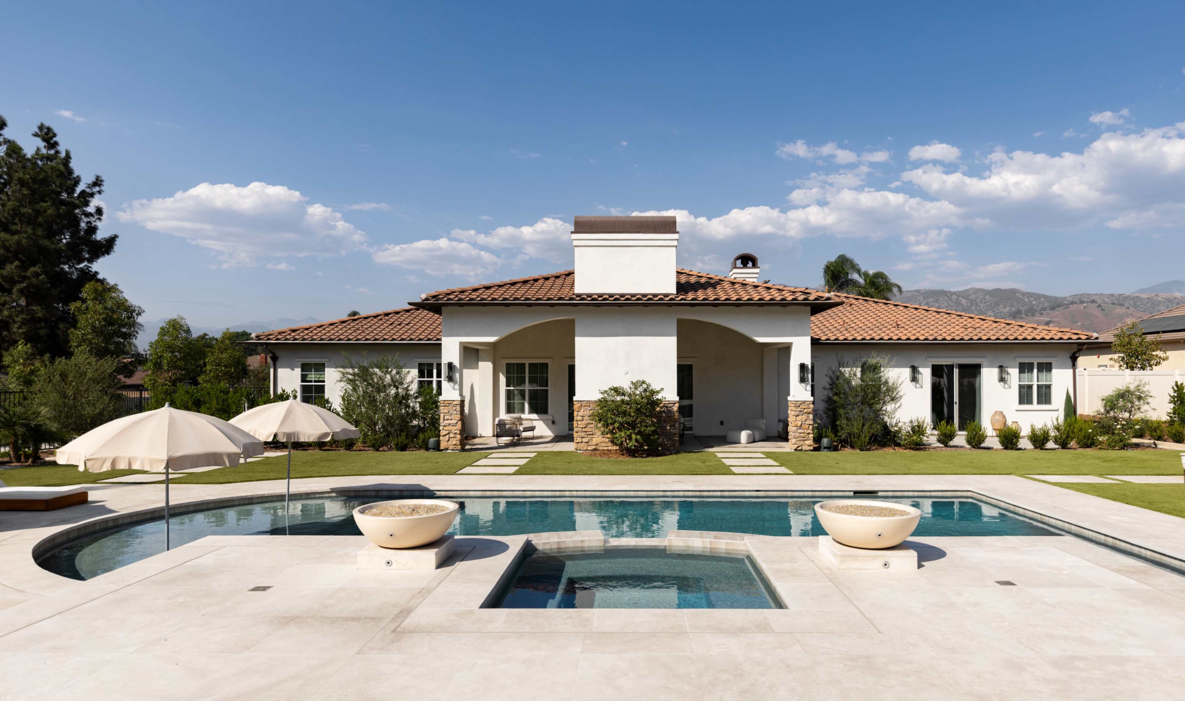 A modern single-story house with a tile roof, surrounded by a landscaped yard and featuring a swimming pool with a spa area in the foreground.