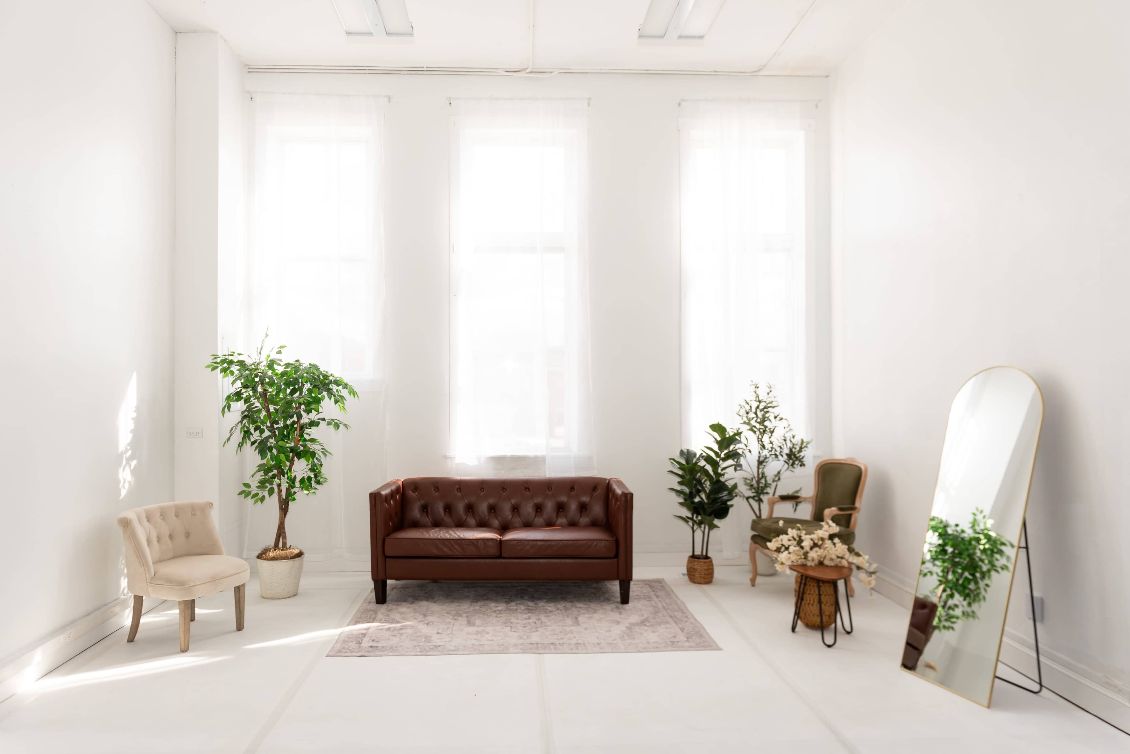 A spacious, brightly lit room features a brown leather sofa, two plants, a decorative mirror, and a small round table with flowers.