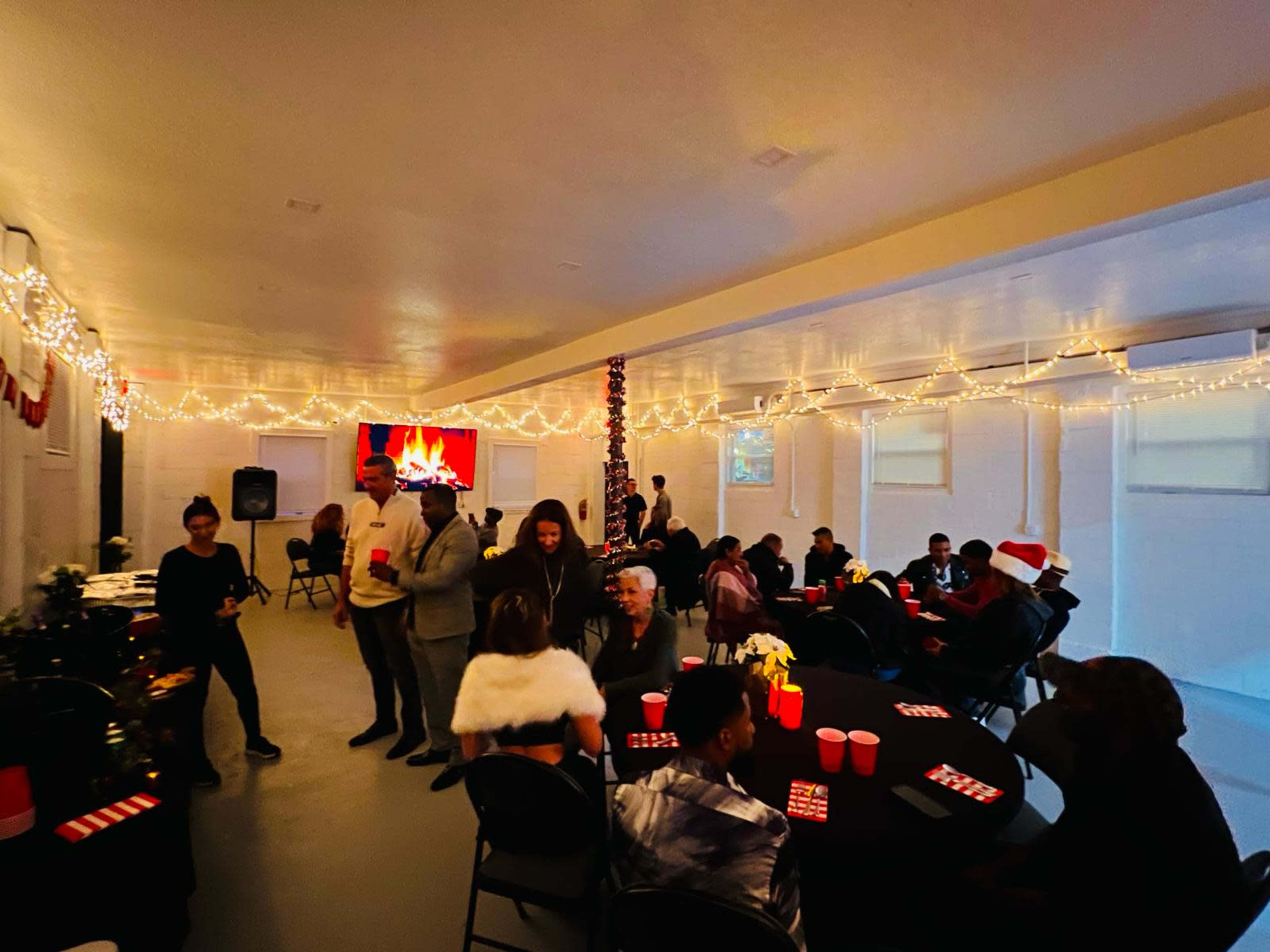 A decorated indoor venue filled with people socializing around tables, featuring festive lighting and a screen displaying a fireplace image.