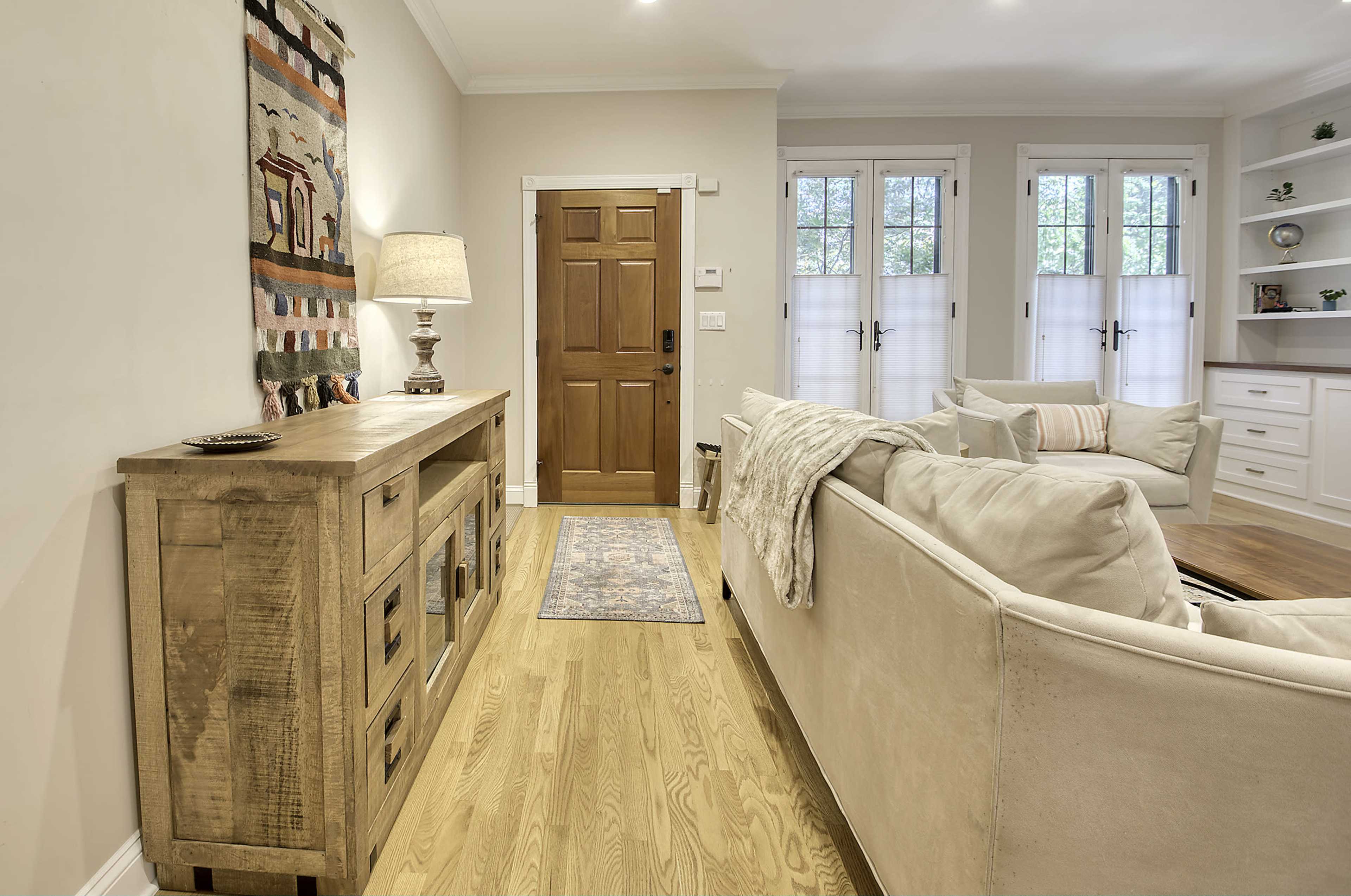 The image shows a well-lit living room featuring a beige sofa, a wooden console table, a lamp, and a wooden front door, with large windows allowing natural light to enter.