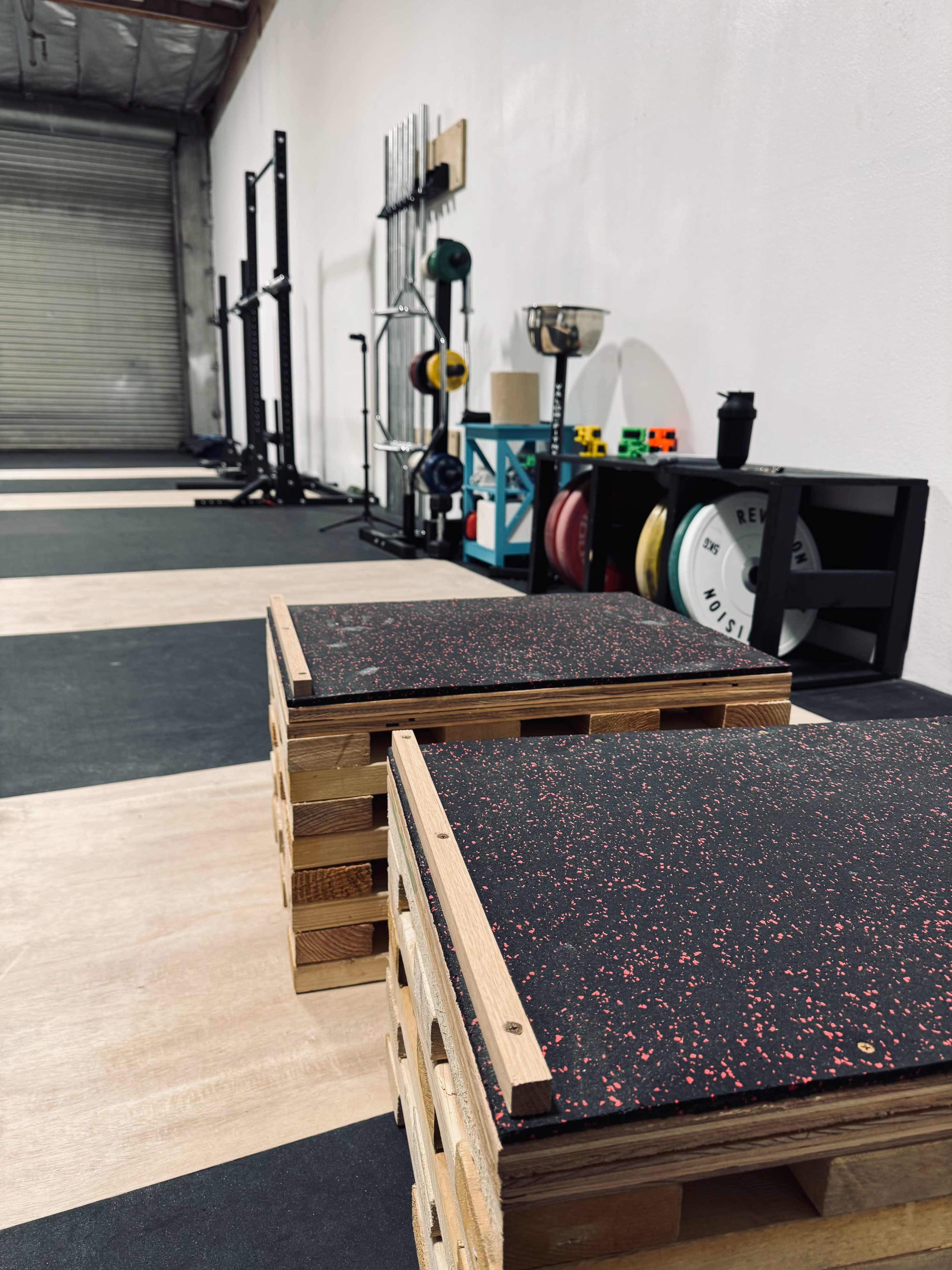 The image shows two wooden plyometric boxes with textured surfaces positioned on a gym floor, surrounded by various exercise equipment such as weights and a pull-up bar in a spacious training area.