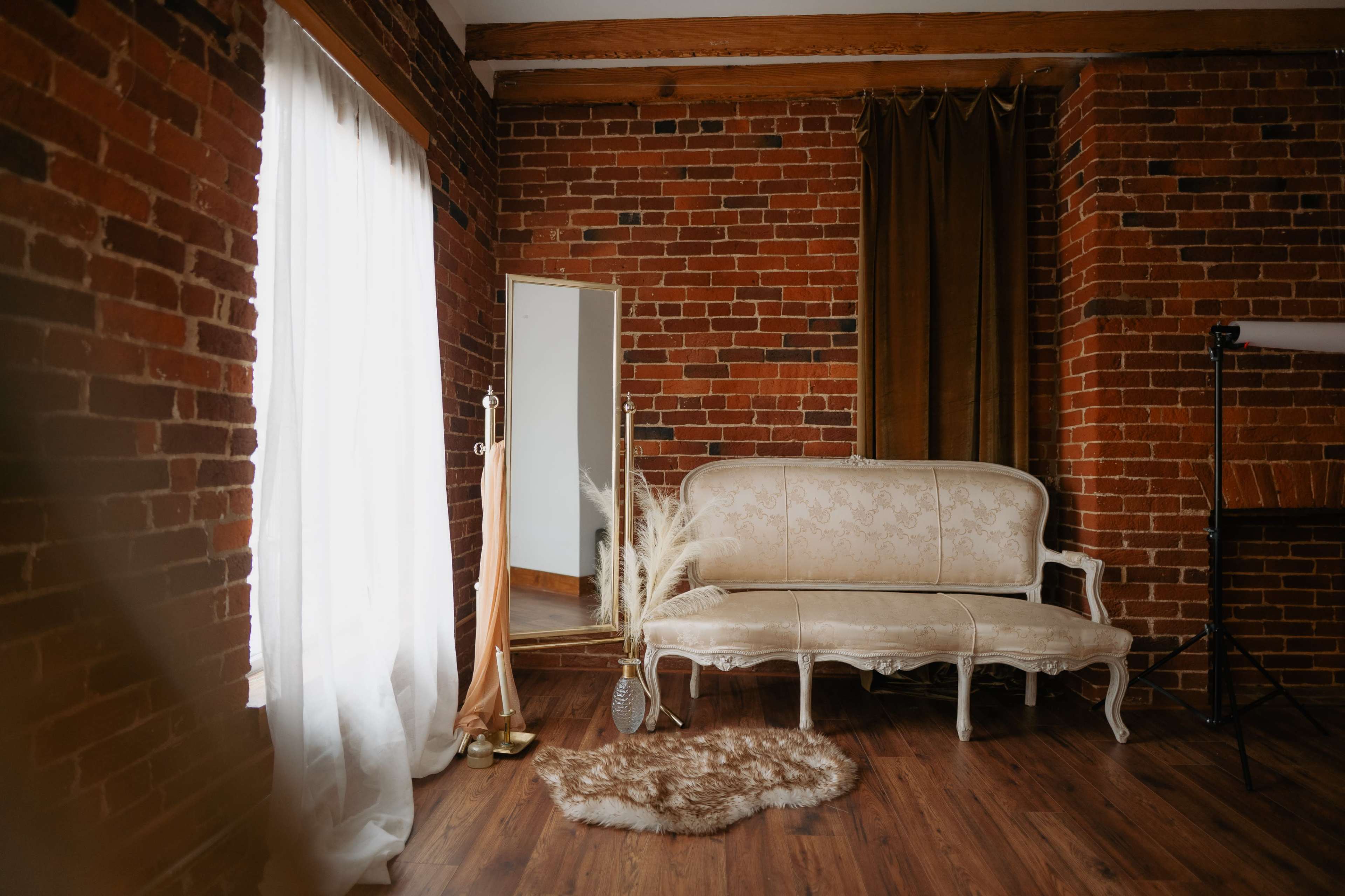 The image features a cozy interior with exposed brick walls, a vintage white sofa, a full-length mirror, and decorative elements like a fuzzy rug and pampas grass.