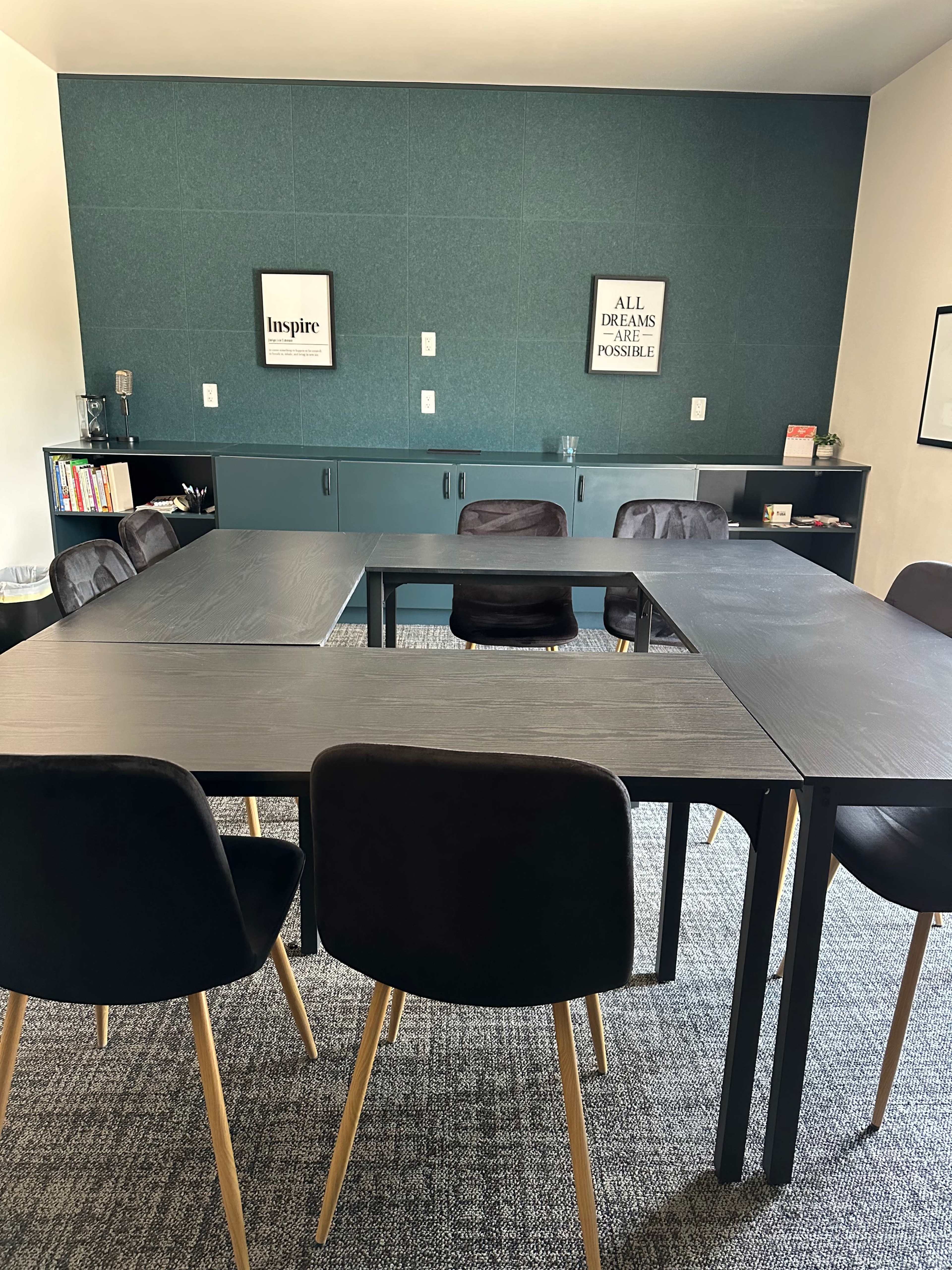 A conference room features a large black table surrounded by black chairs, with a green accent wall displaying framed artwork.