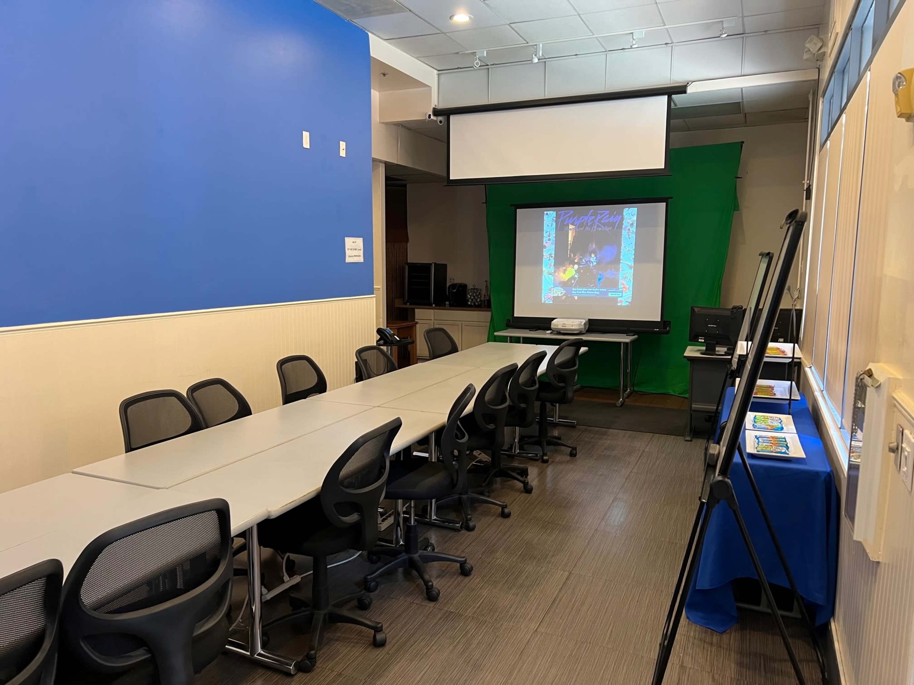 A conference room features a long table with chairs, a projector screen against a green backdrop, and a display table with promotional materials.
