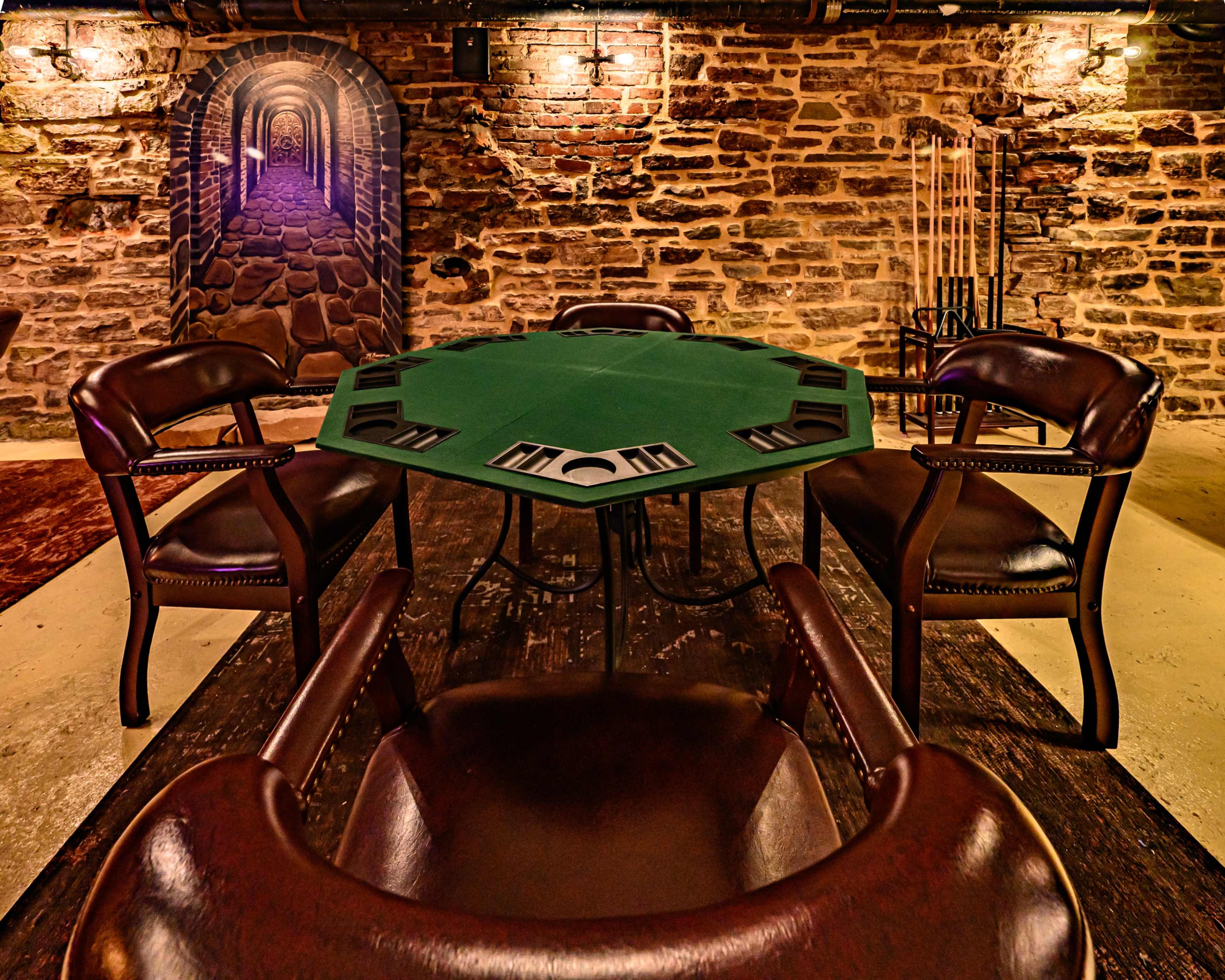 A poker table with a green felt surface is surrounded by four brown leather chairs in a dimly lit stone-walled basement.