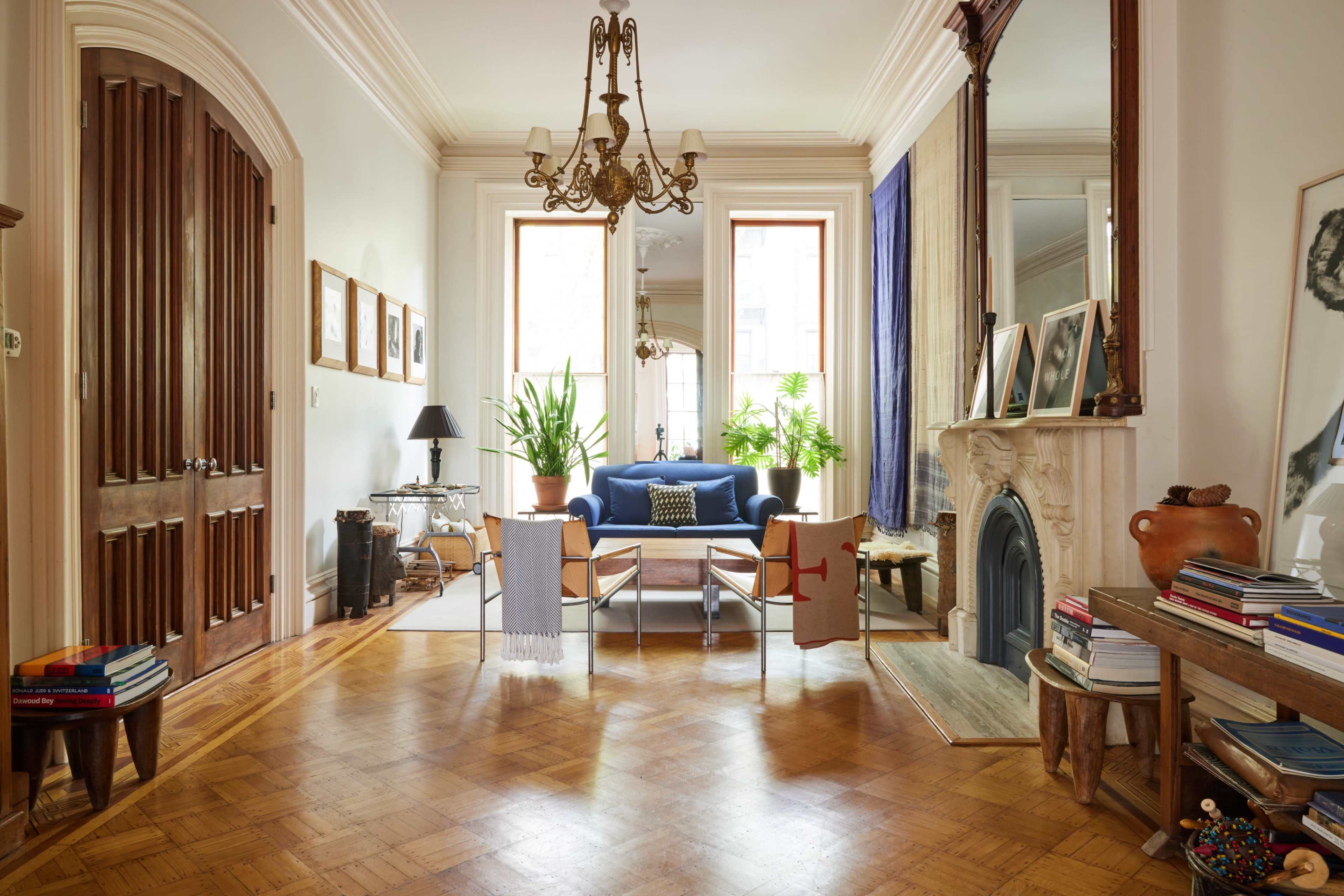 The image shows a well-lit living room with a blue sofa, a coffee table, plants, and a decorative fireplace, featuring wooden doors and a large mirror on the walls.