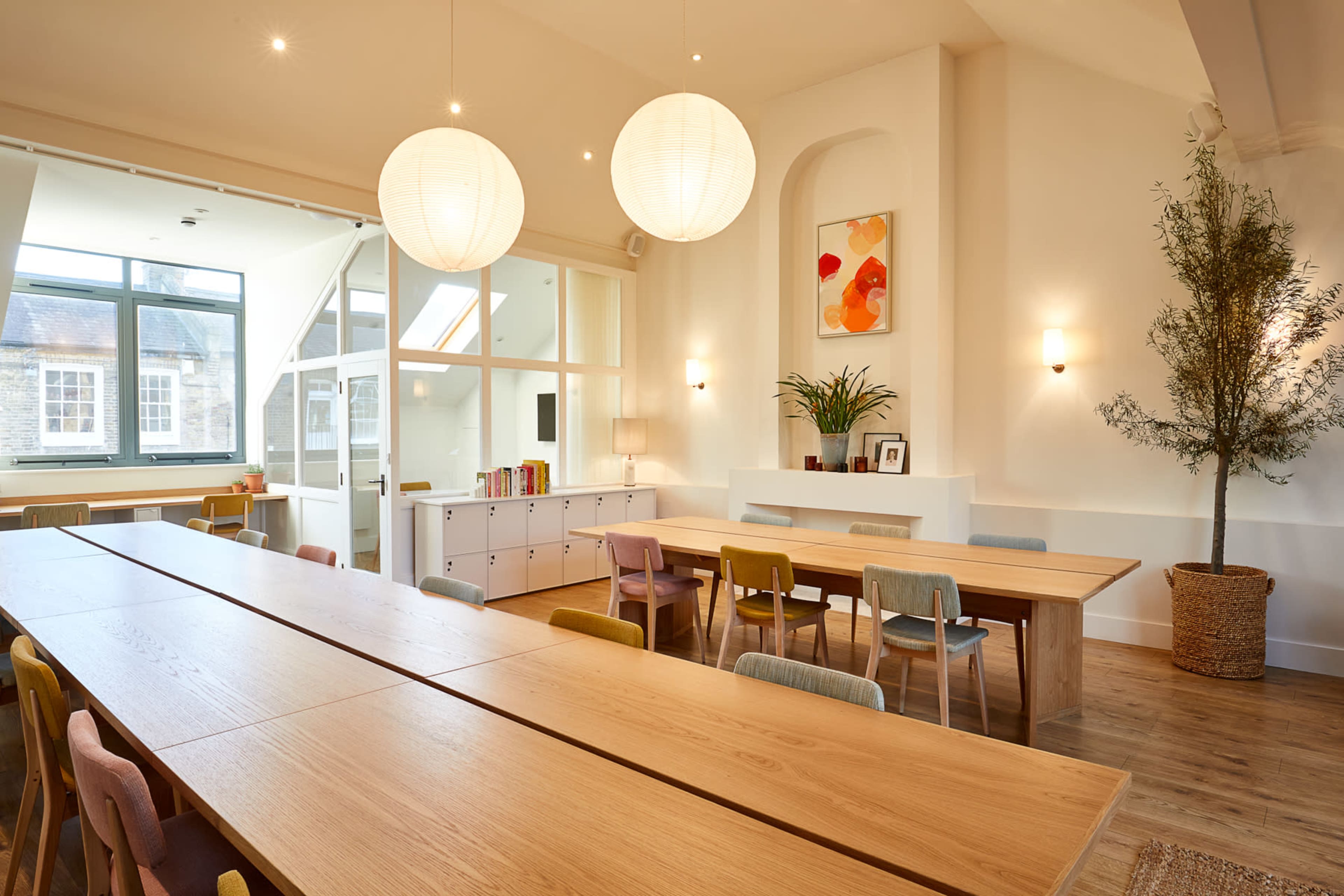 A spacious, well-lit room features long wooden tables with colorful chairs, potted plants, and decorative artwork on the walls.