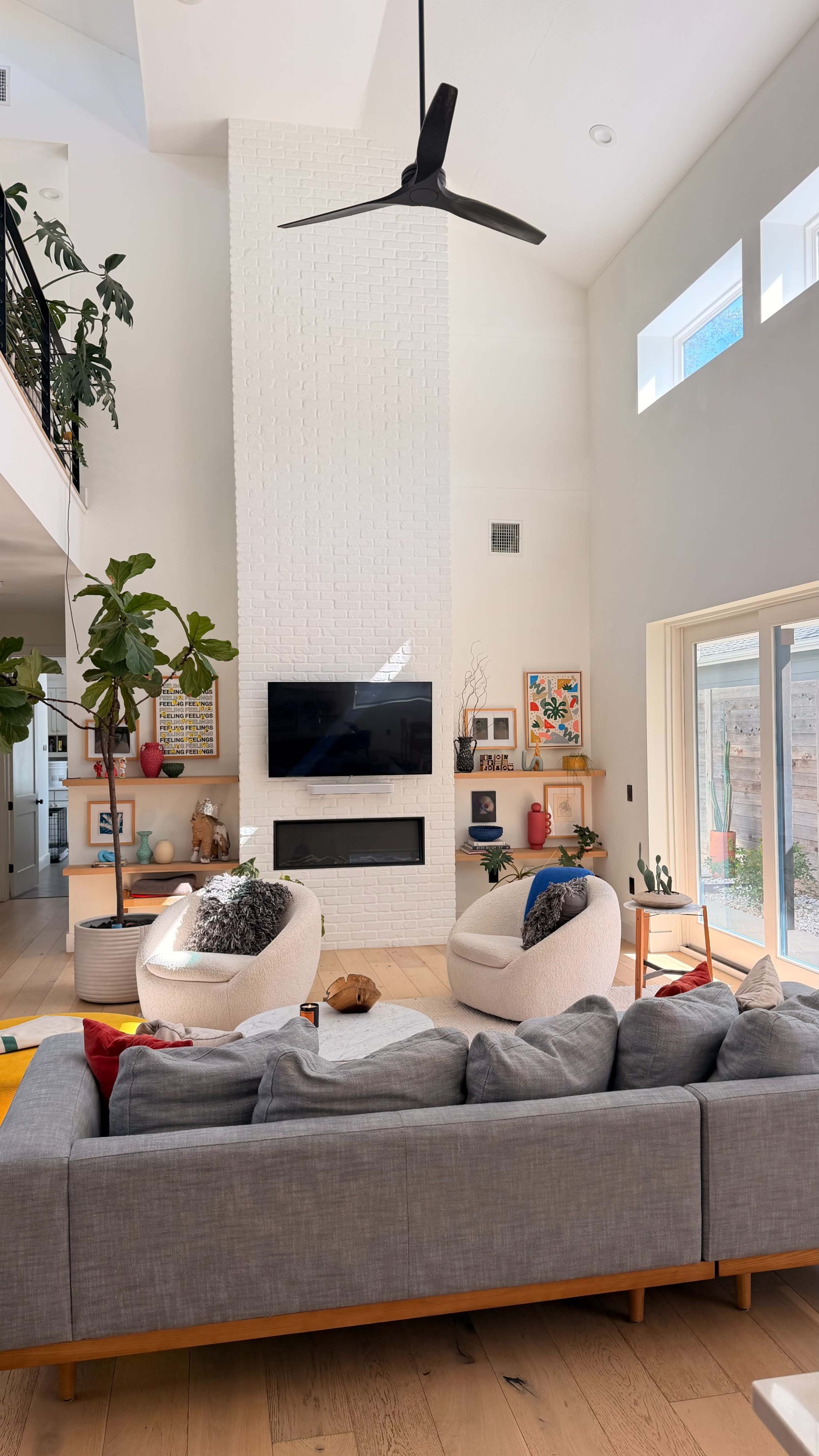 The image showcases a spacious, modern living room with a large sectional sofa, two round armchairs, a tall indoor plant, and a flat-screen TV mounted on a white brick wall.