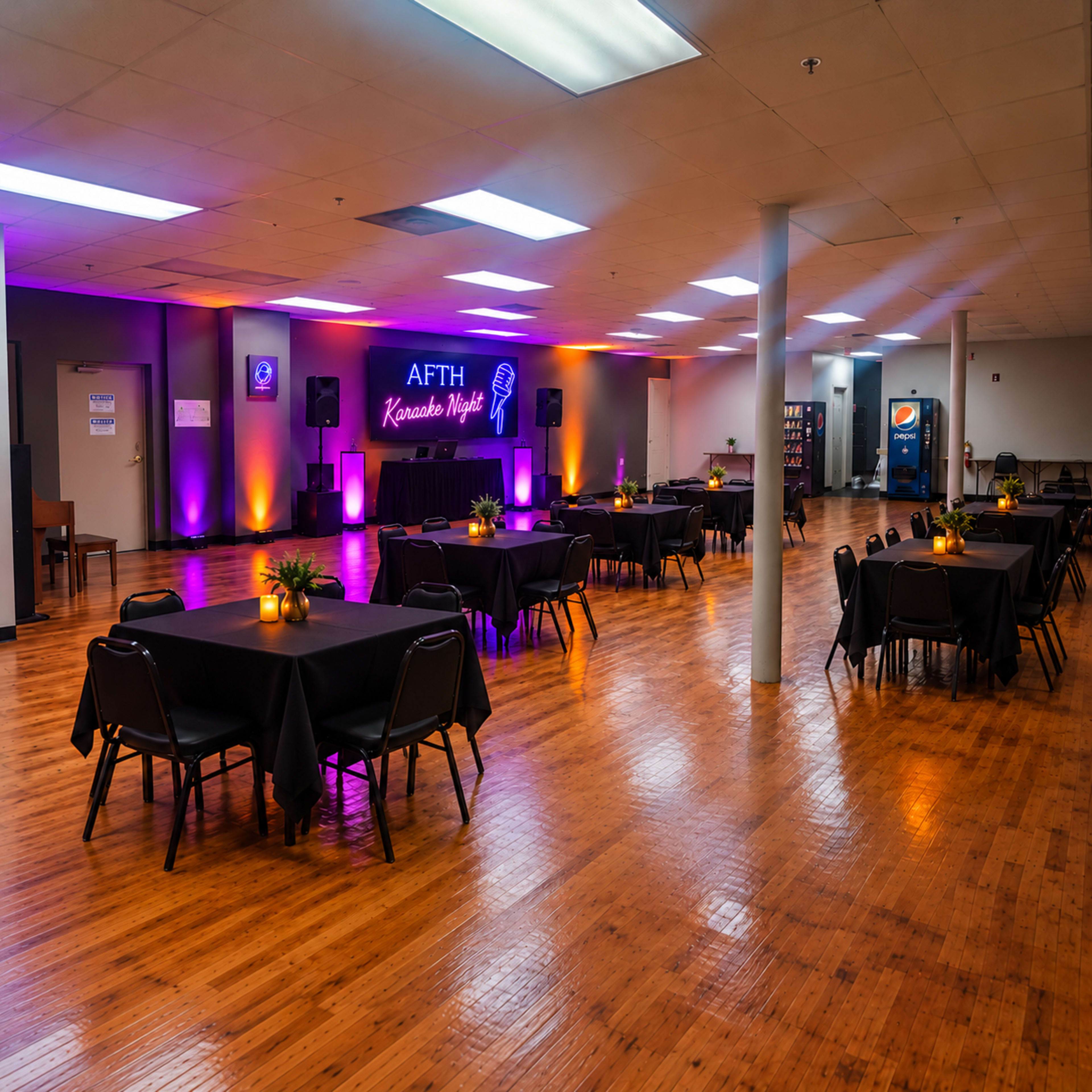 The image shows a spacious event room arranged with several black tables and chairs, illuminated by purple and orange lights, featuring a stage for karaoke at one end.