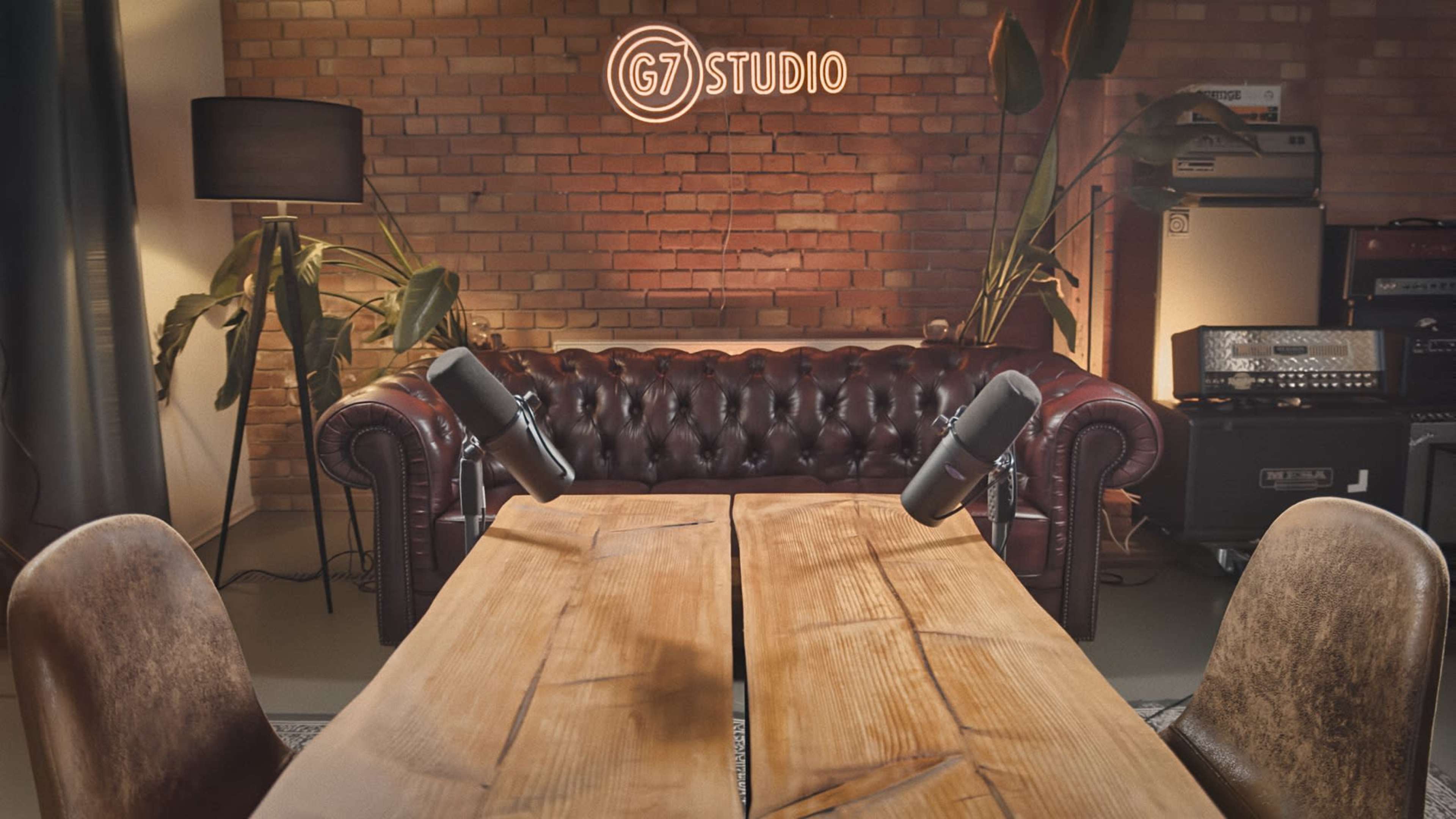 The image shows a recording studio with a brown leather couch, wooden table, and two microphones set up in front of a wall with a glowing "G7 STUDIO" sign.