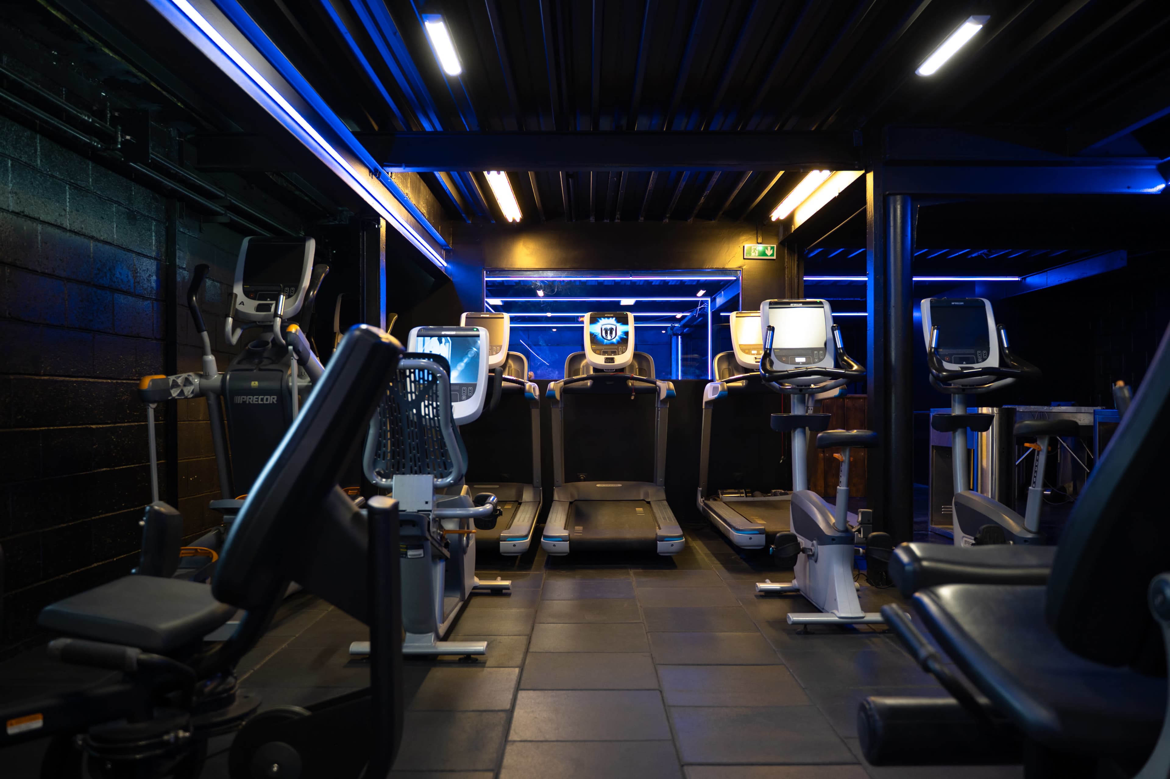 The image shows a dimly lit gym space featuring various exercise machines along the walls and illuminated by blue LED lights.
