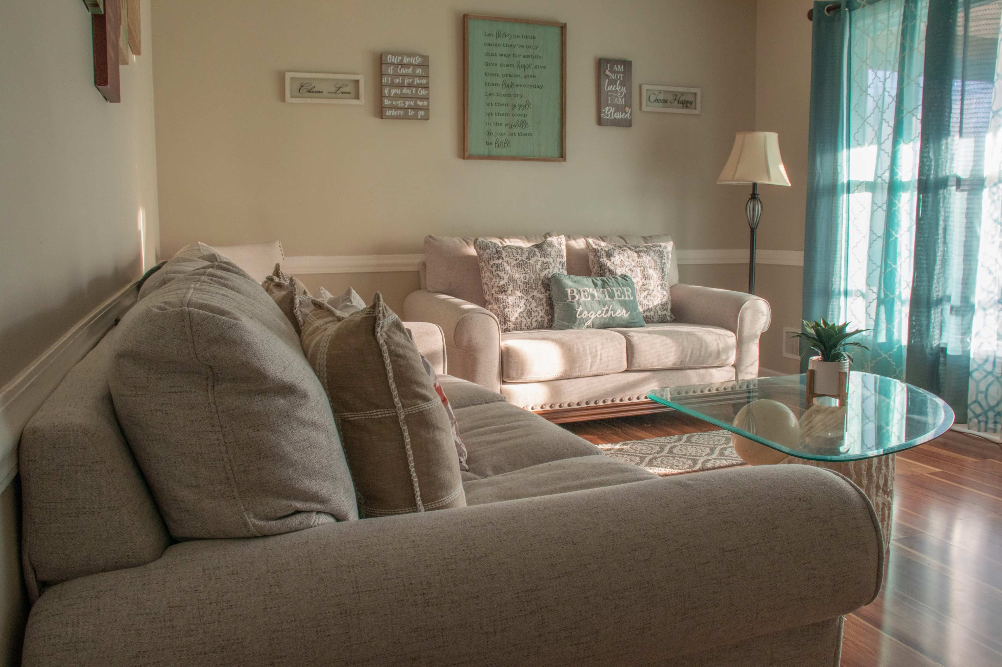 A living room features a light-colored sofa and armchair, a glass coffee table, and decorative wall art, with sunlight streaming through a window with sheer curtains.