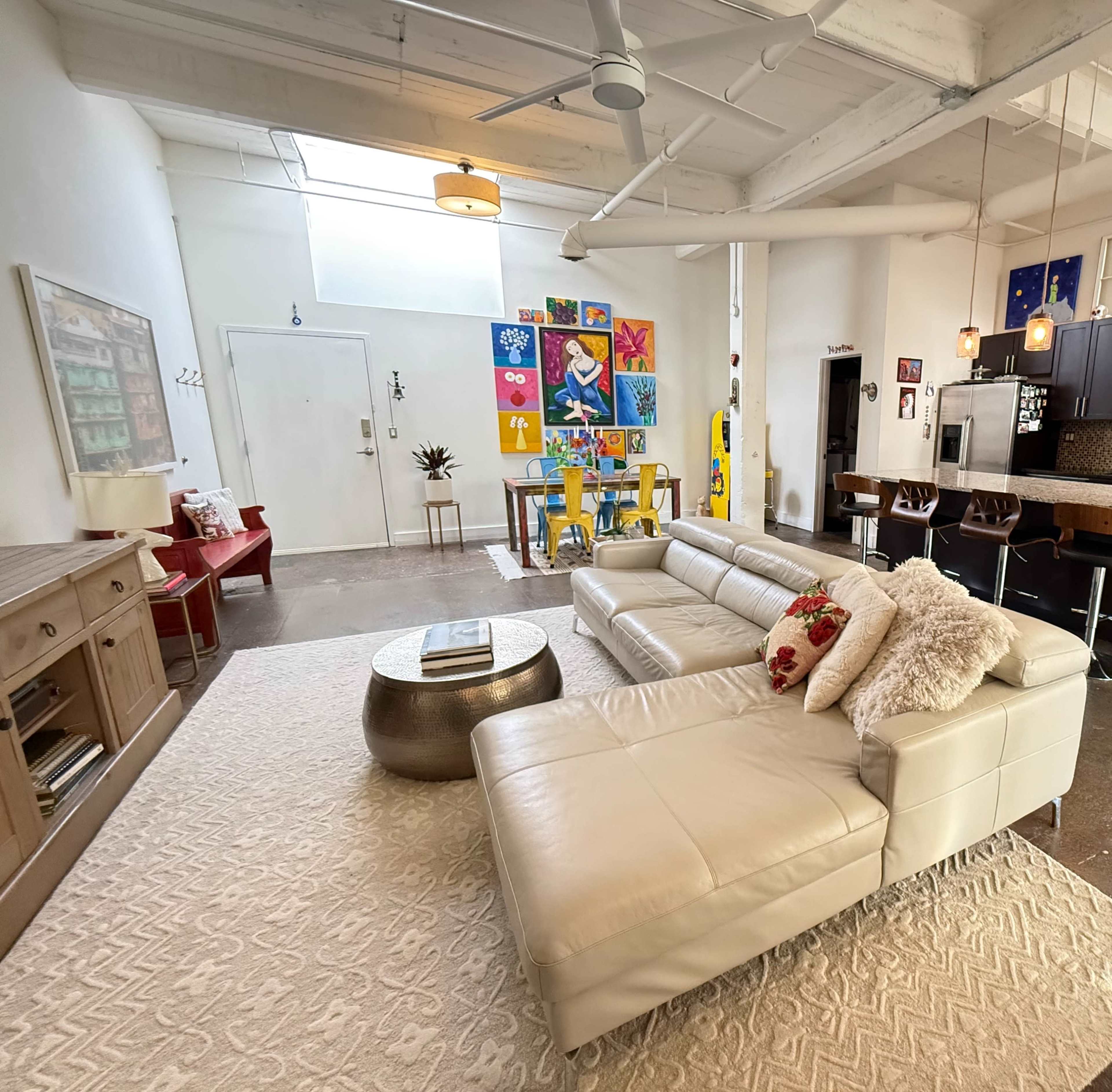 The image shows a modern living room with a light-colored sectional sofa, a round coffee table, a variety of colorful artwork on the wall, and a kitchen area in the background.