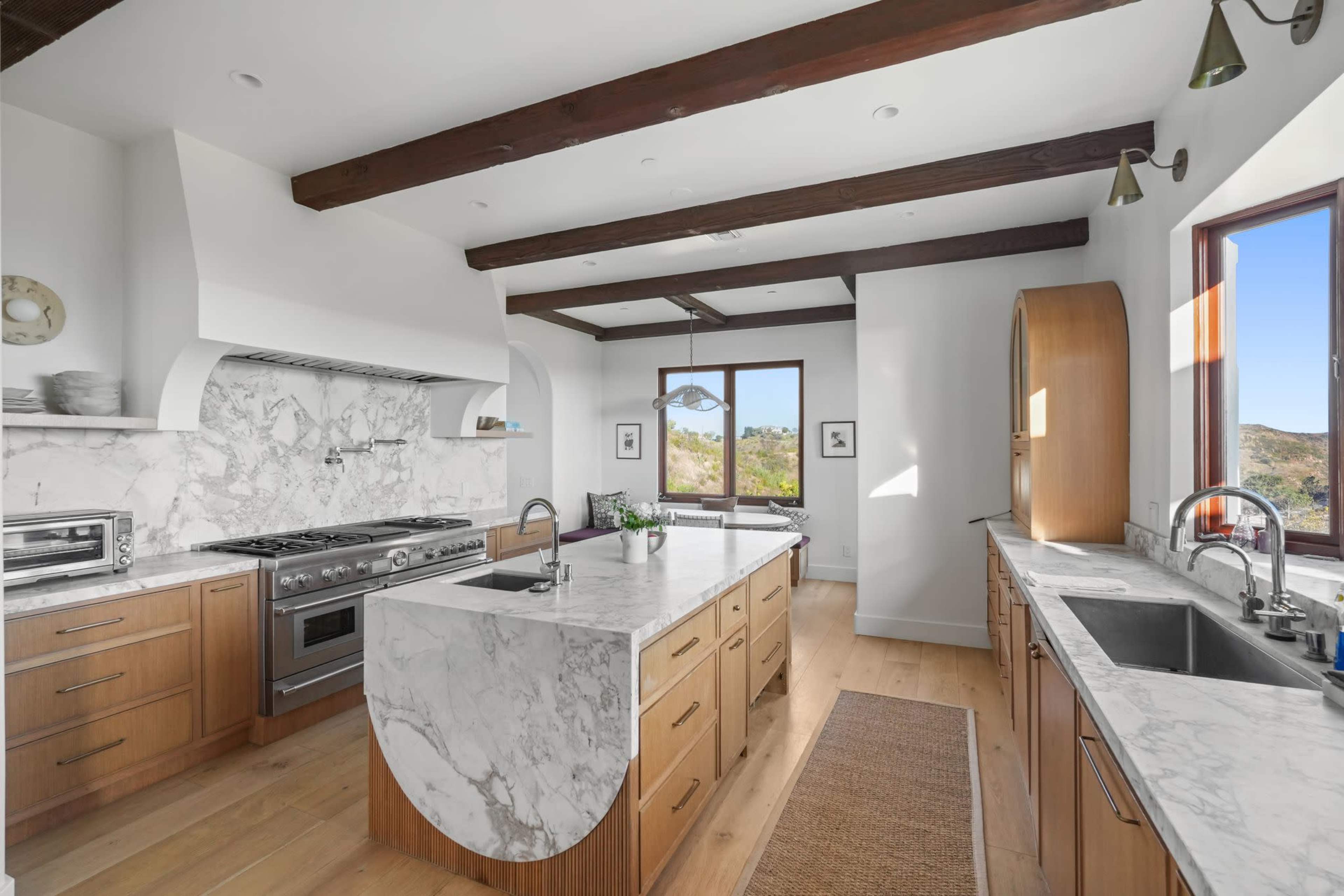 The image shows a modern kitchen with marble countertops, wooden cabinetry, and a large island, featuring large windows that provide a view of the outdoors.