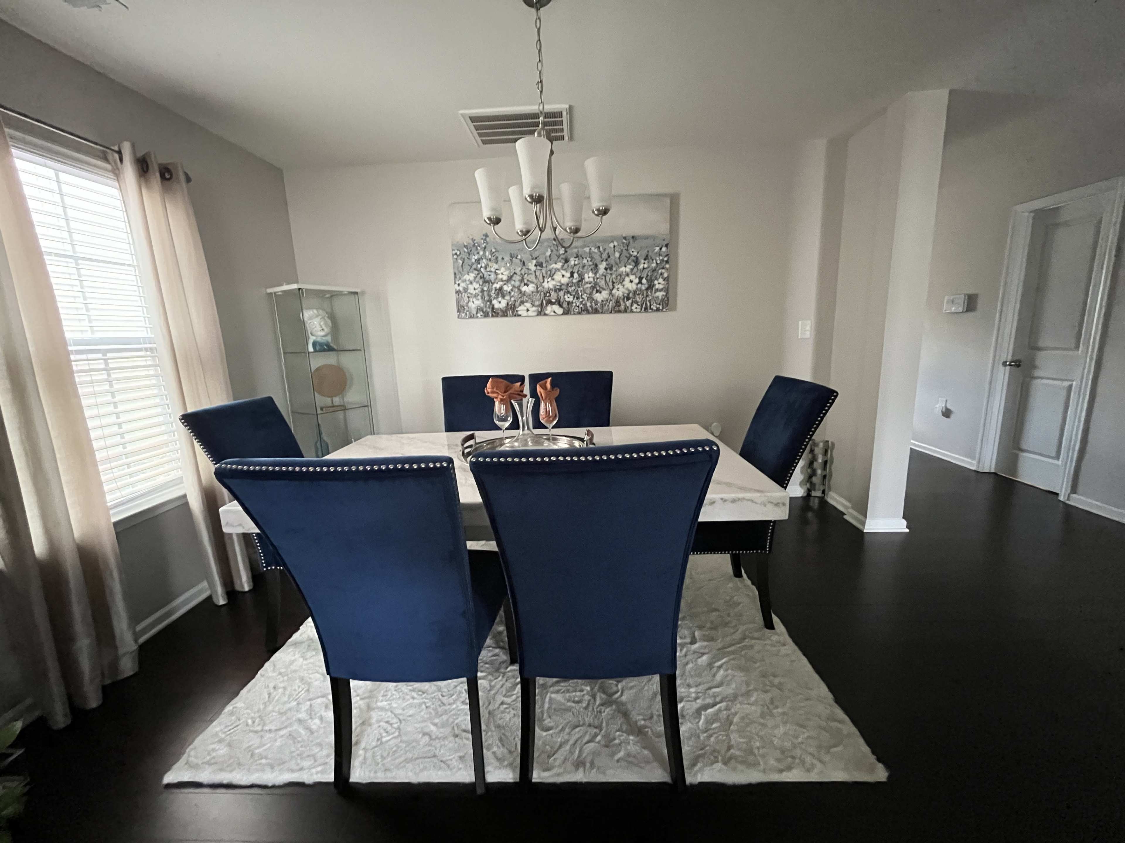 A dining room features a rectangular table surrounded by four blue upholstered chairs, with a chandelier overhead and a decorative painting on the wall.