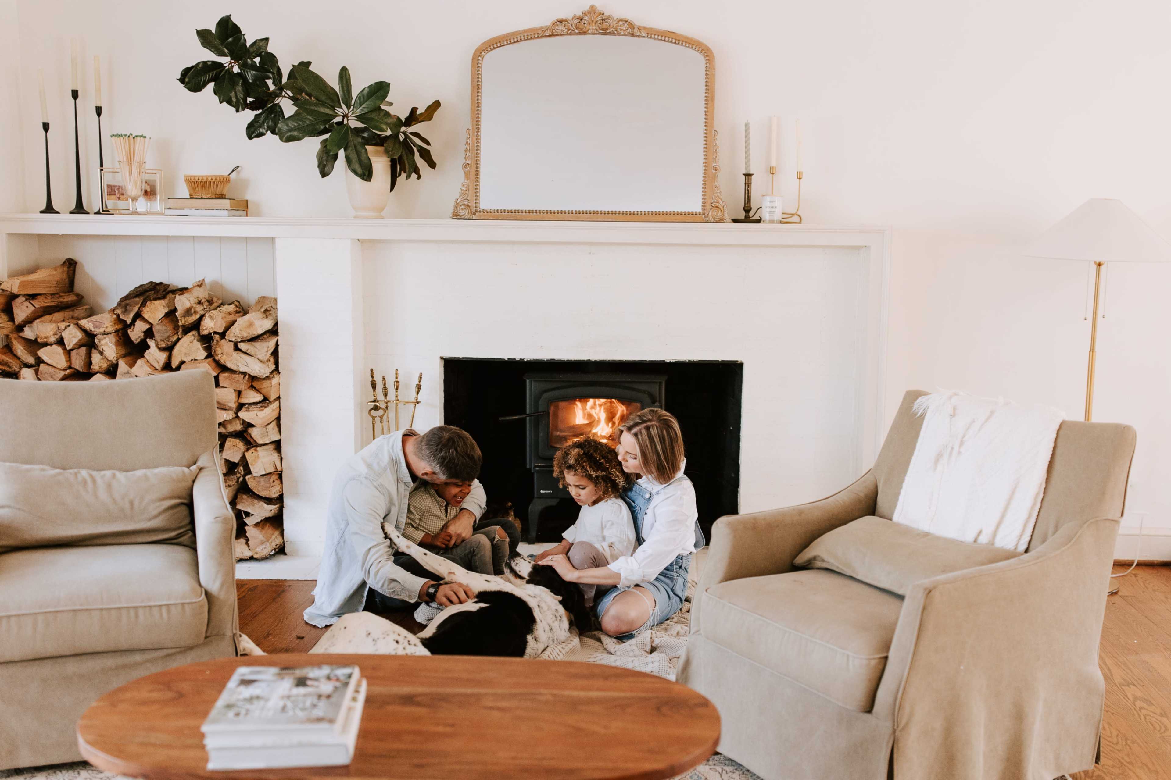 A family of four sits on the floor in front of a fireplace, engaged in an activity together.