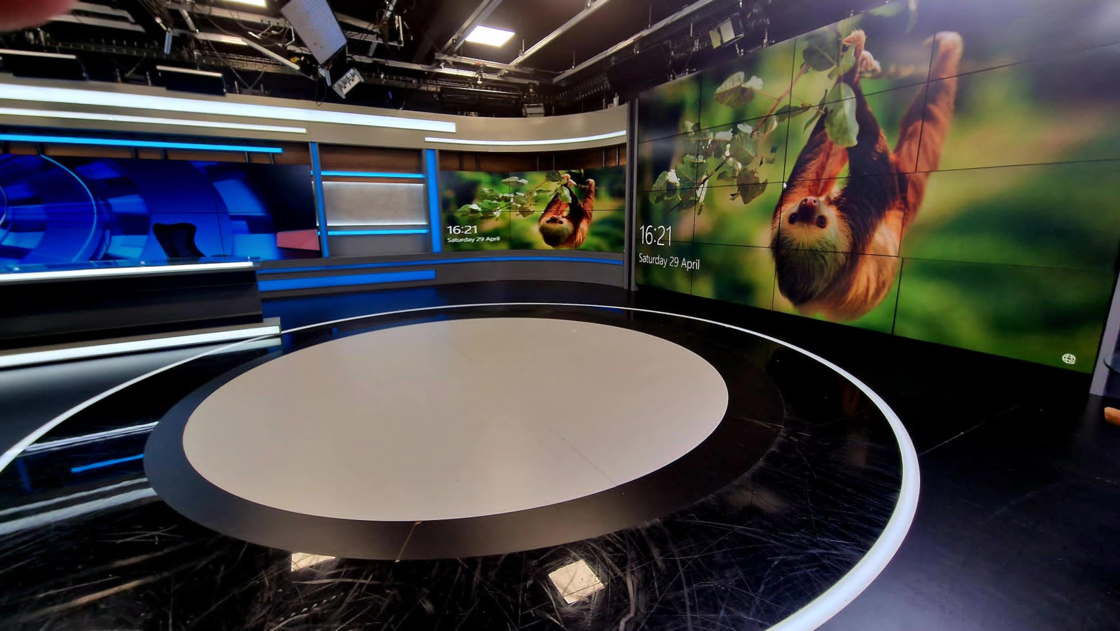 The image shows a modern television news studio with a large circular stage and high-definition screens displaying a sloth and the time.