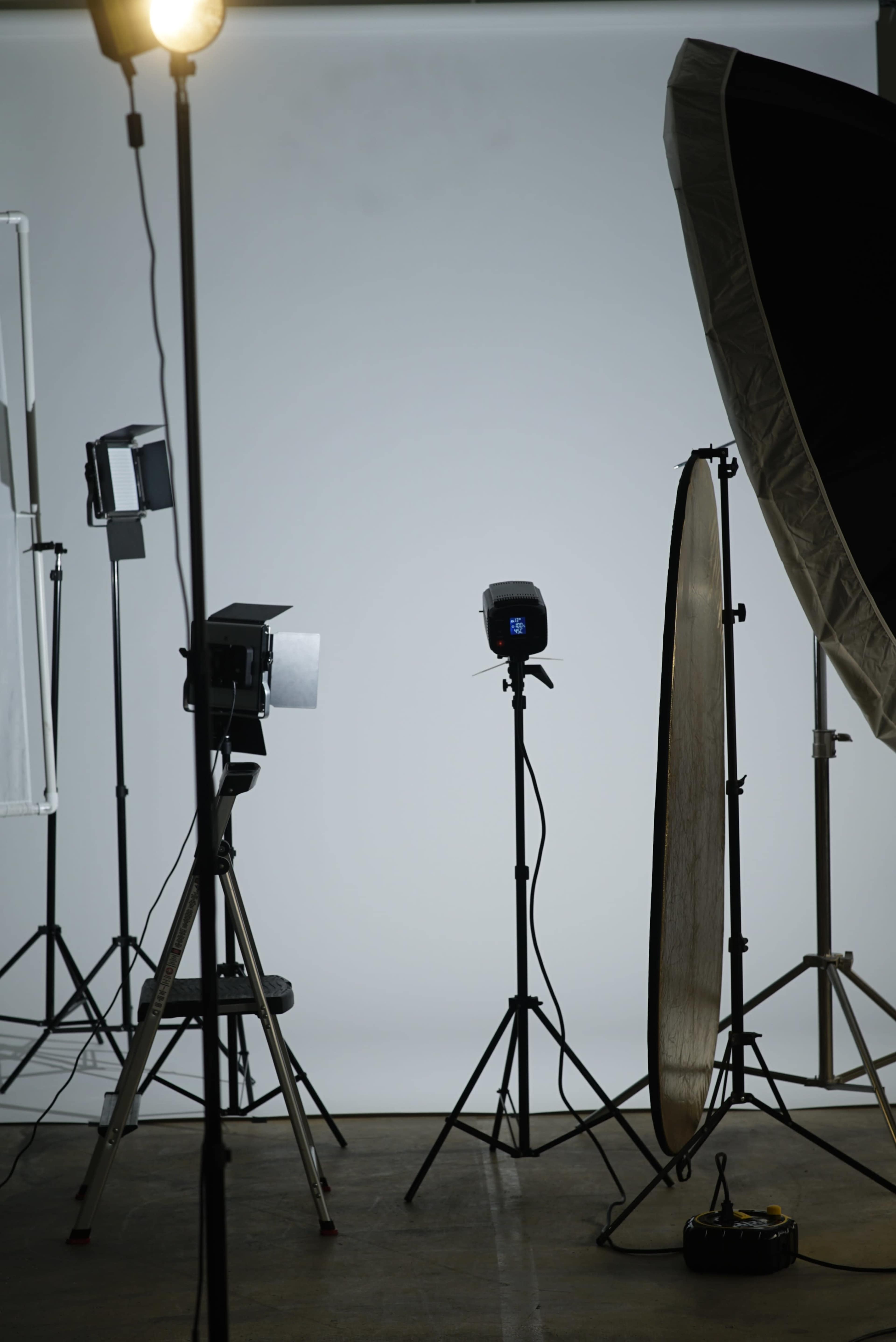 The image shows a photography studio setup with various lighting equipment, including softboxes and reflectors, arranged on tripods against a neutral background.