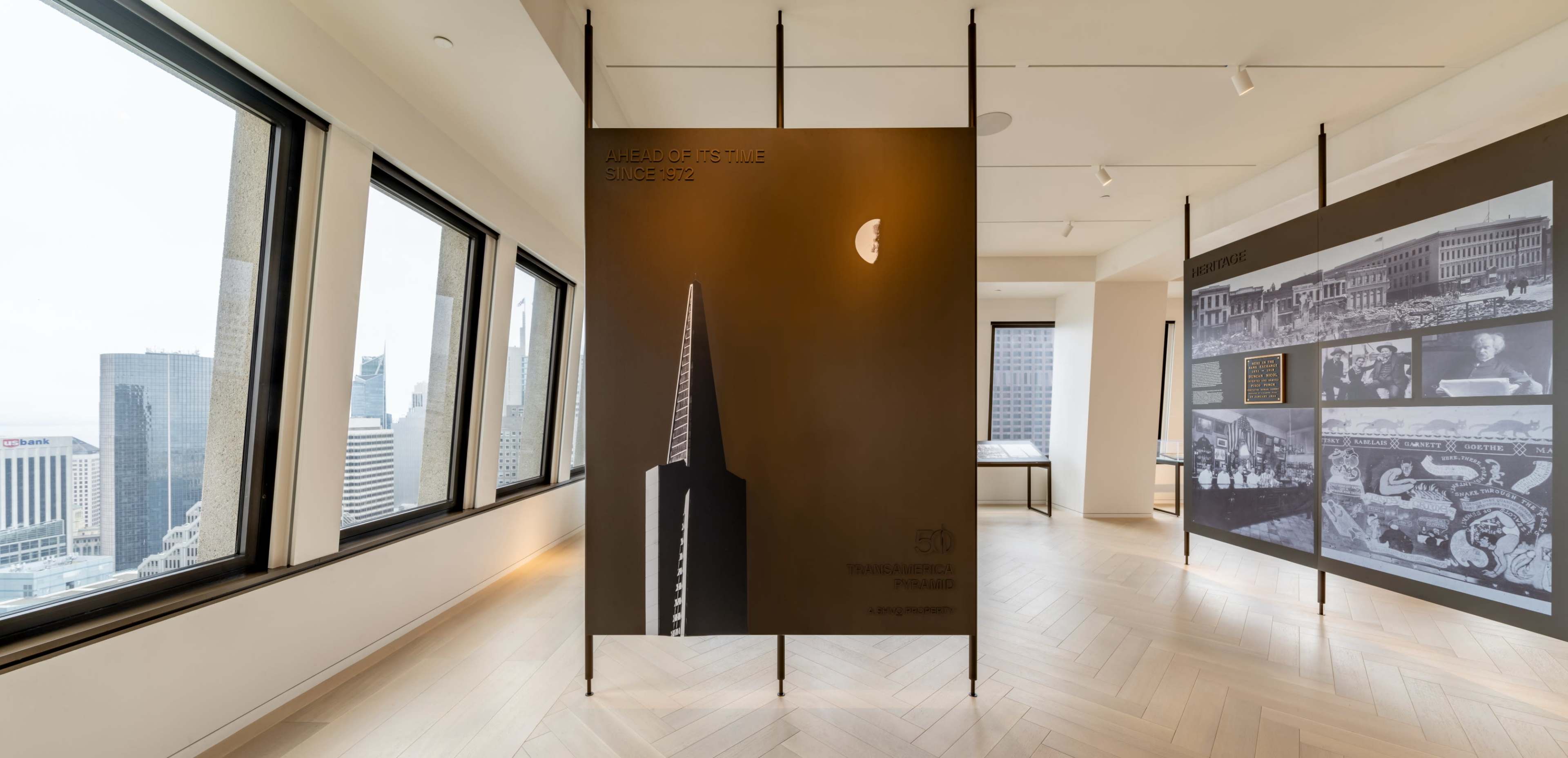 The image shows a modern exhibition space with tall panels displaying information and visuals, surrounded by large windows offering a view of city skyscrapers.