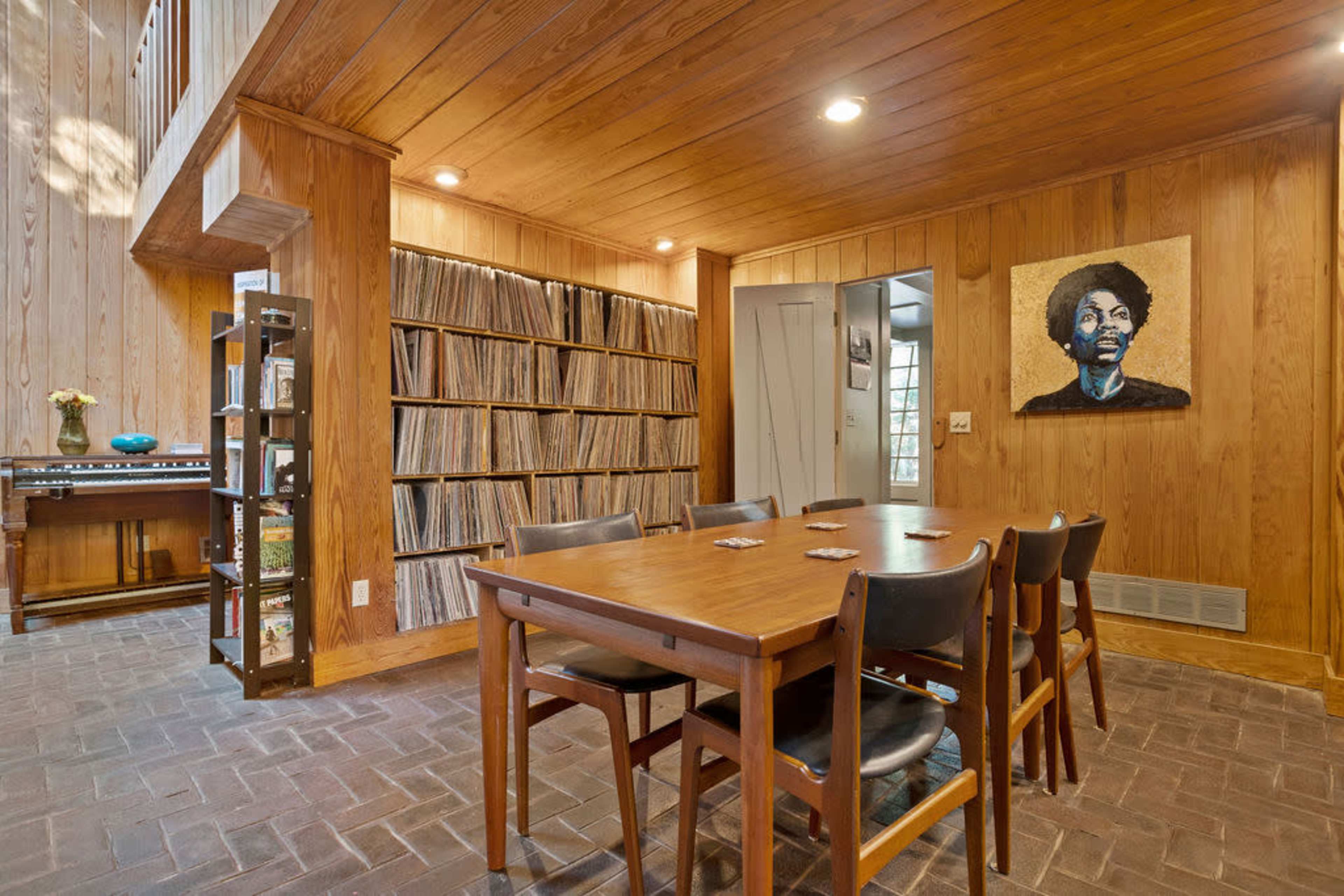 A wooden-floored room features a dining table surrounded by chairs, a wall of vinyl records, and a portrait on the wall.