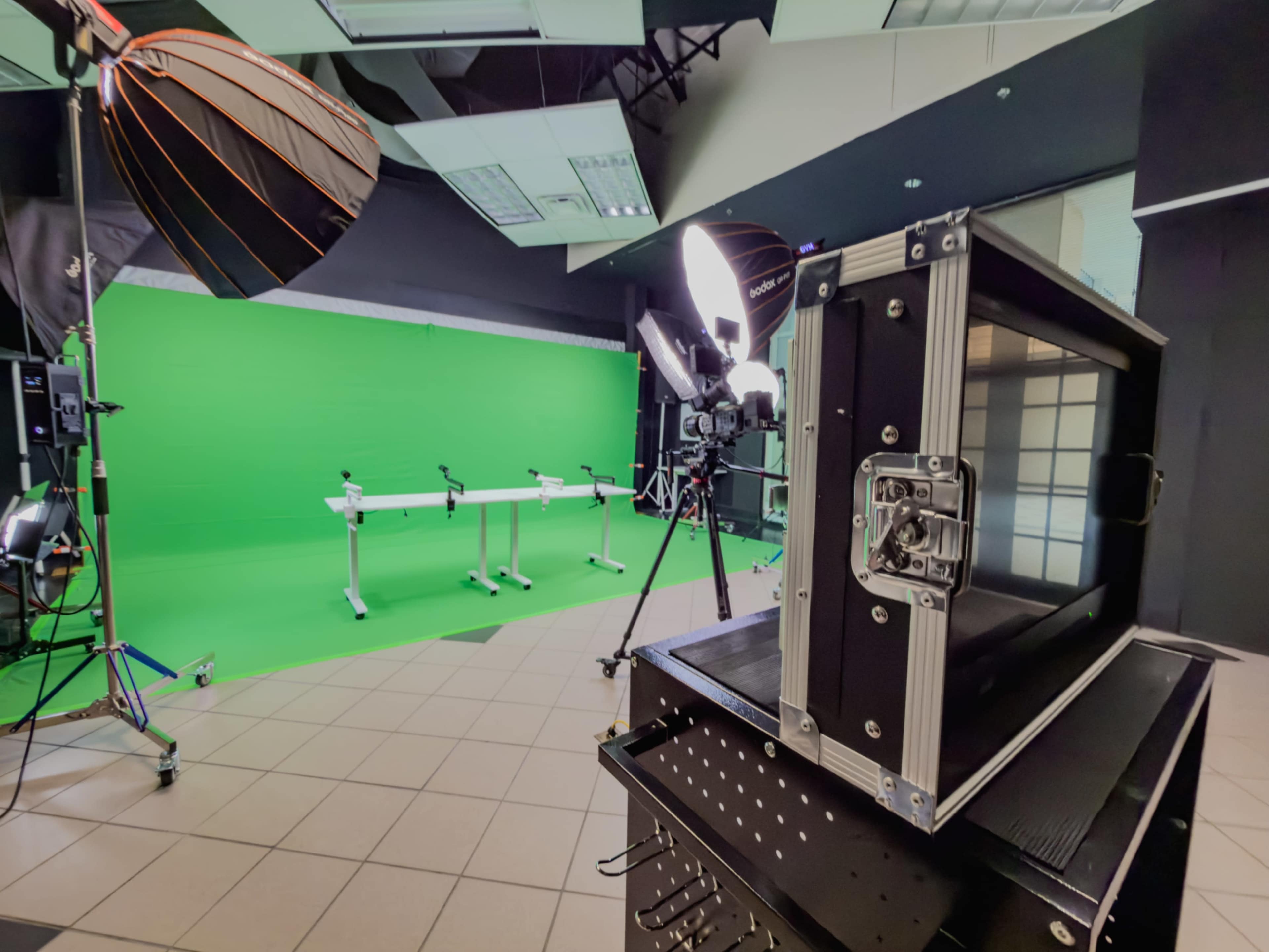The image shows a studio setup featuring a green screen background, lighting equipment, and a camera positioned near a black equipment case.