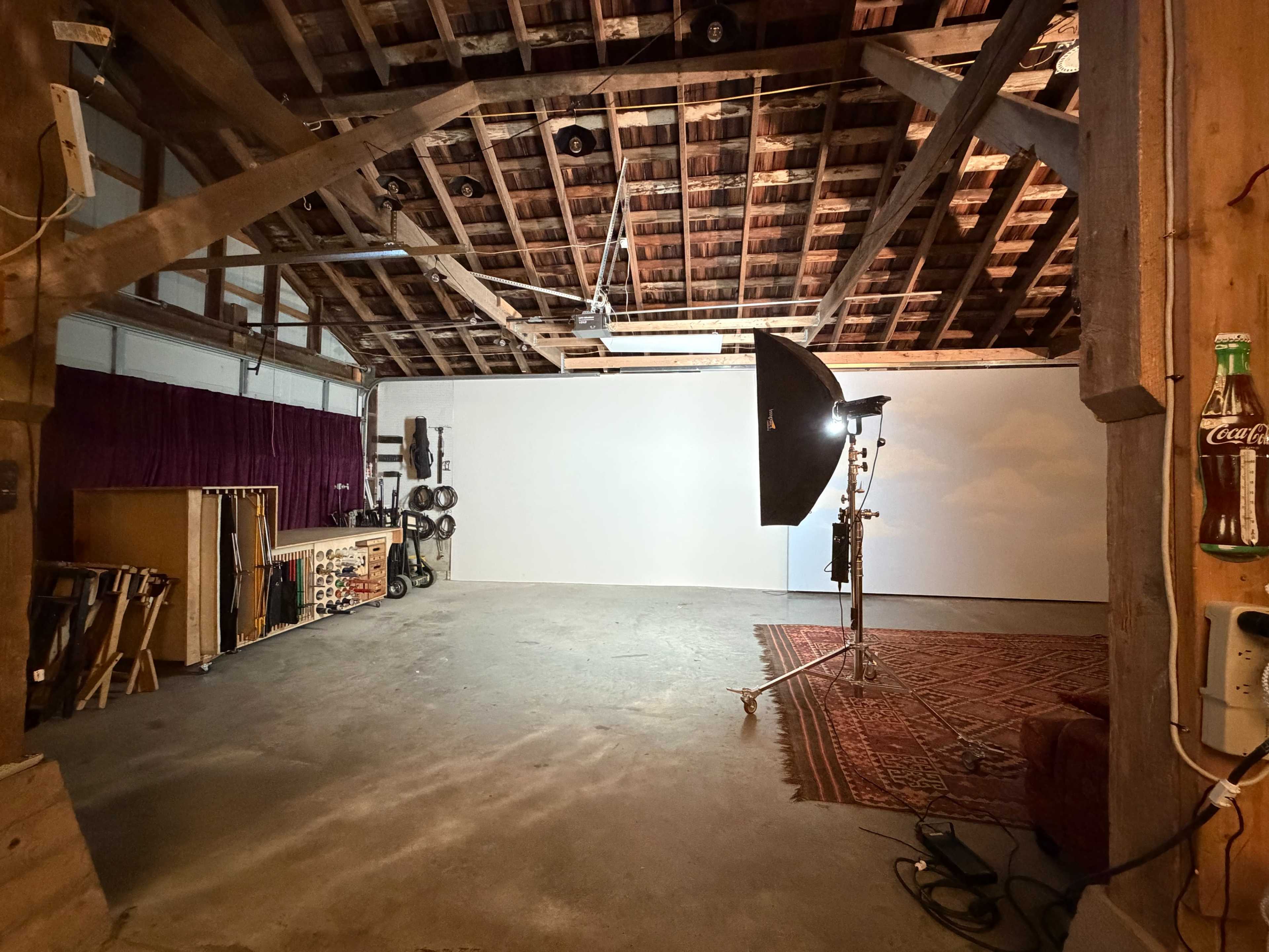 The image shows a spacious, partially furnished studio with exposed wooden beams, a white wall, and a lighting setup on a stand.