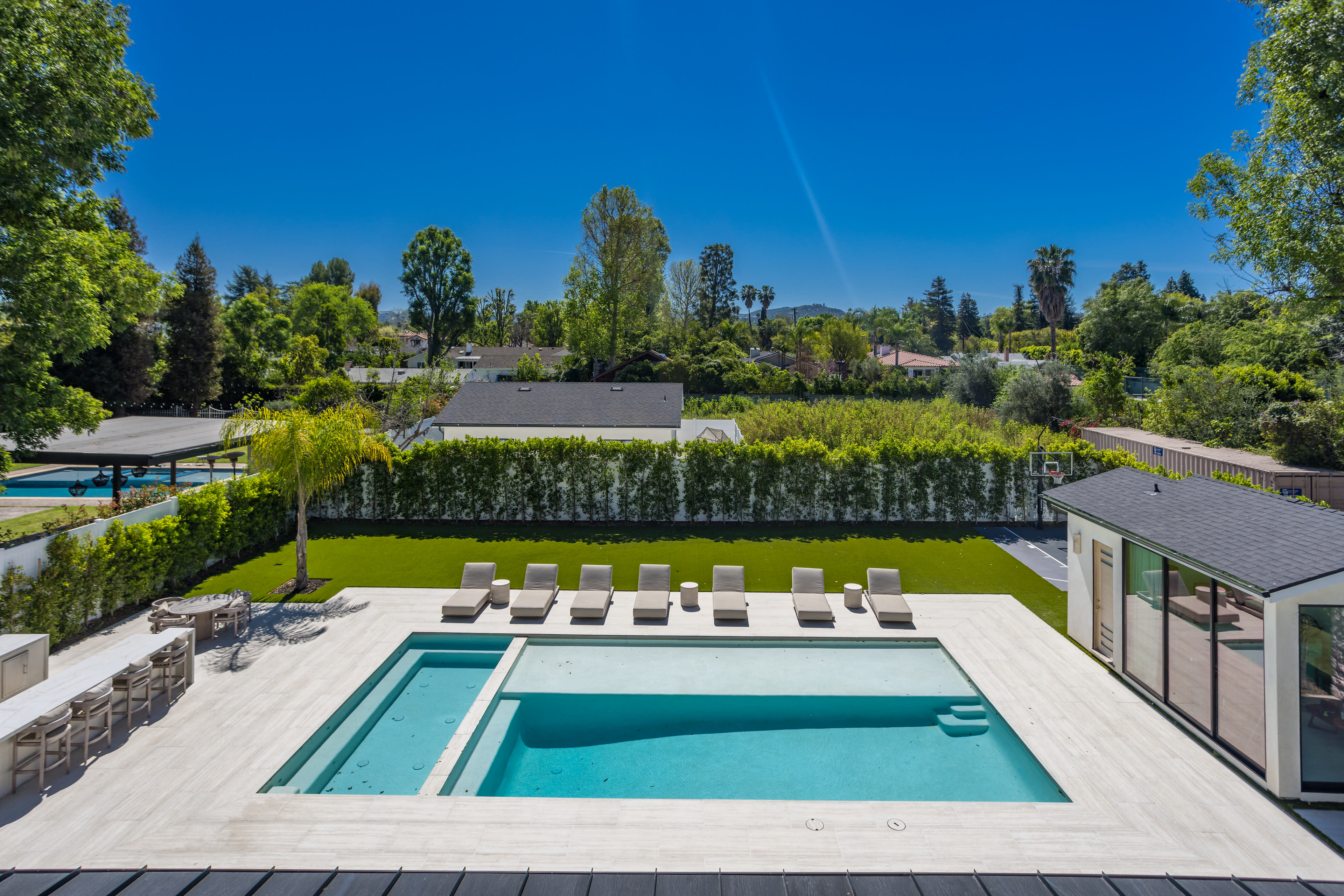 The image shows a modern home with an expansive pool area featuring lounge chairs, surrounded by lush greenery and trees under a clear blue sky.