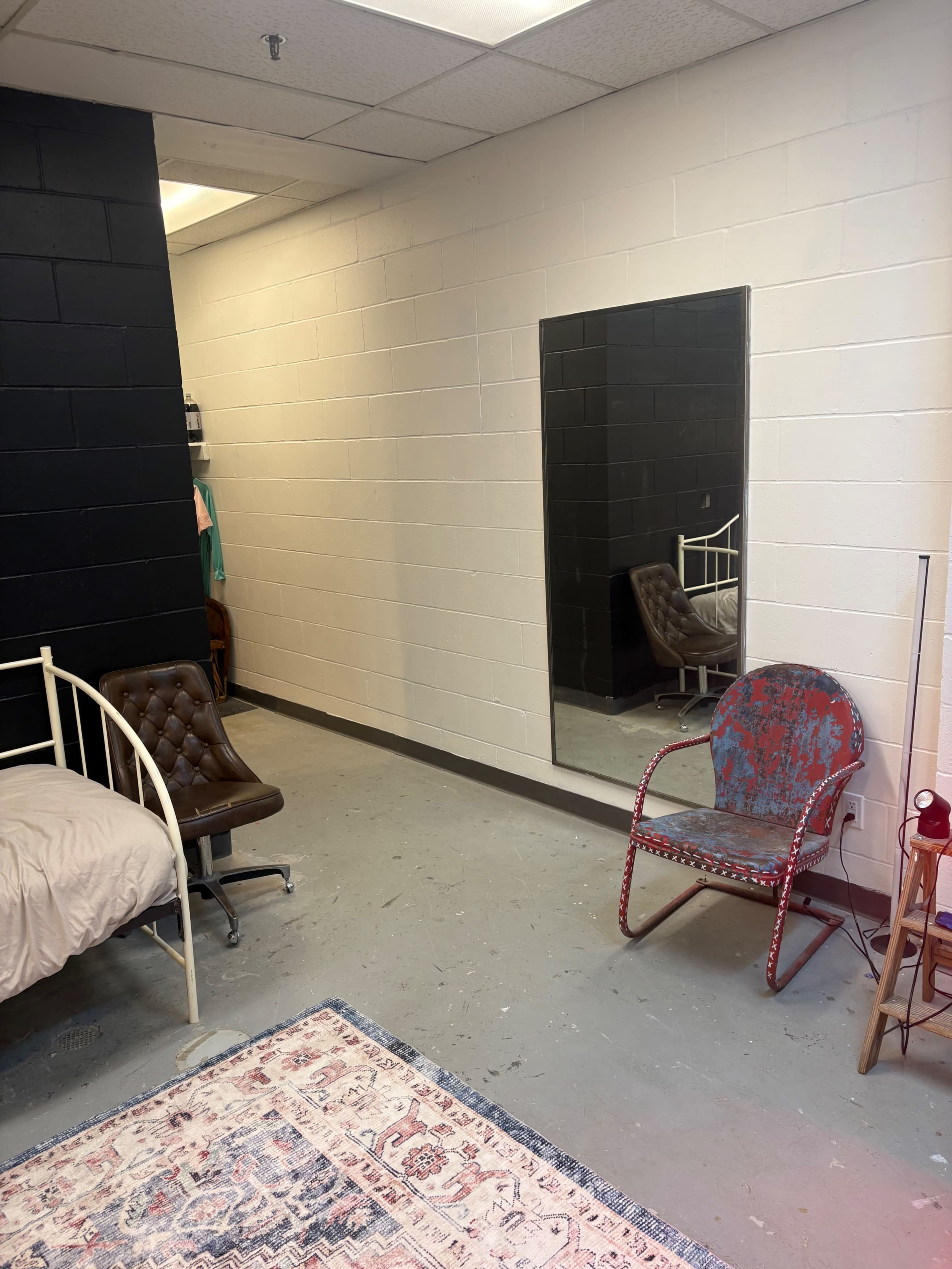 The image shows a sparse room featuring a bed, a chair, a large mirror on the wall, and a patterned rug on the floor.