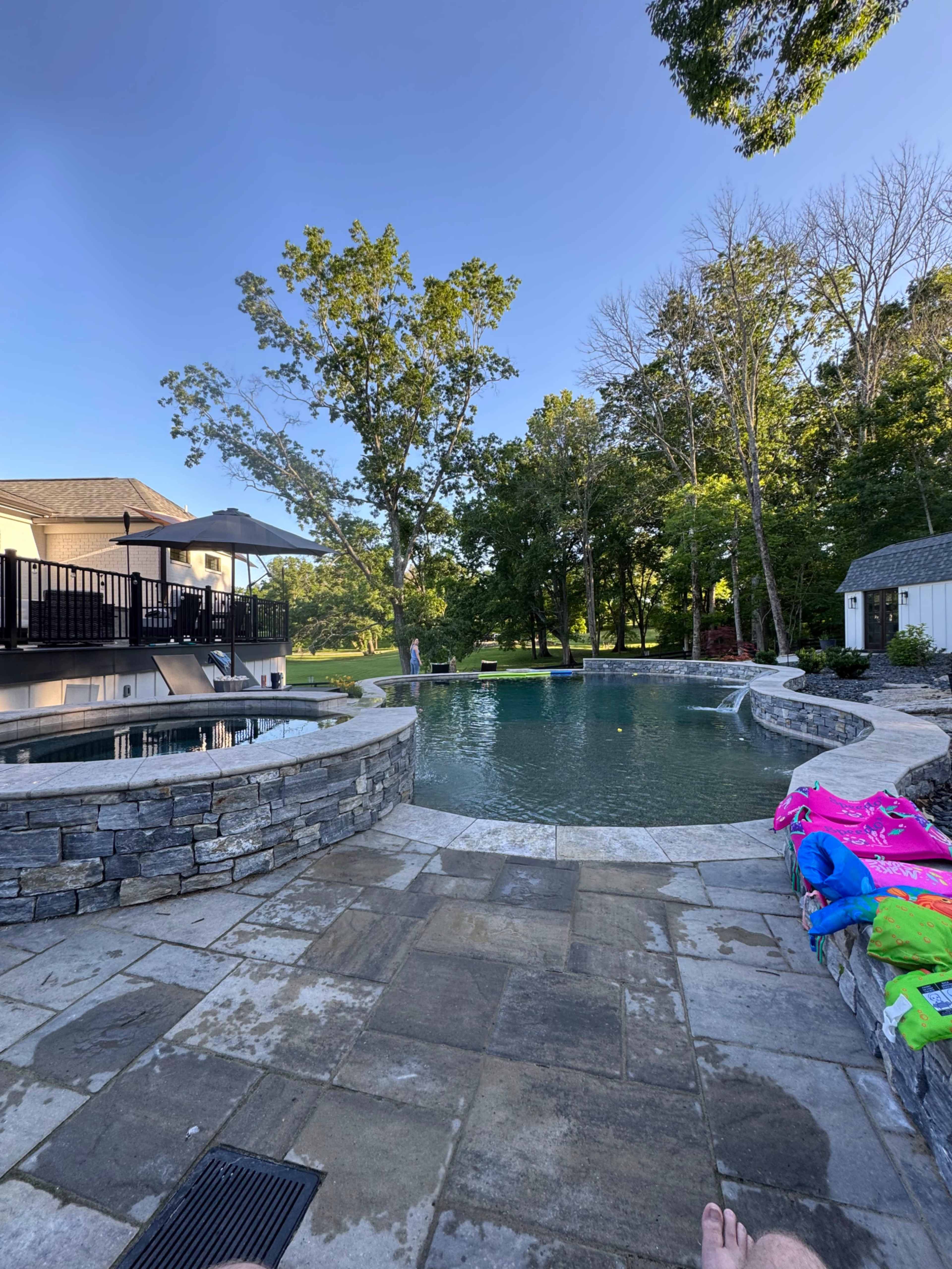 The image shows a landscaped backyard with a stone-lined swimming pool, surrounded by trees and a lawn.