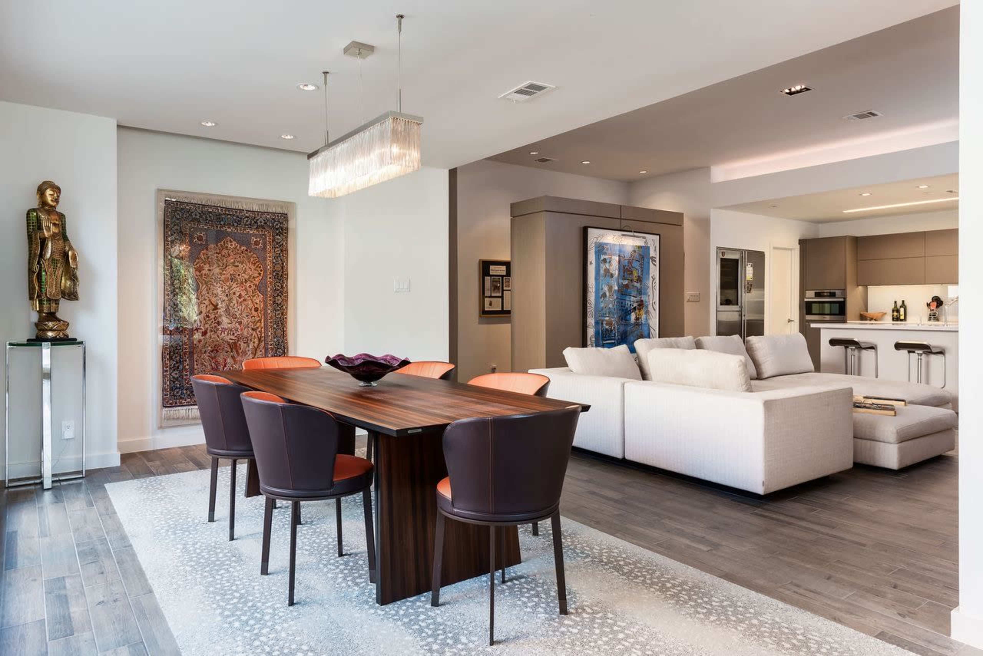 A modern dining area featuring a dark wooden table surrounded by orange and brown chairs, with a decorative rug beneath and an adjacent living space furnished with a light-colored sofa.