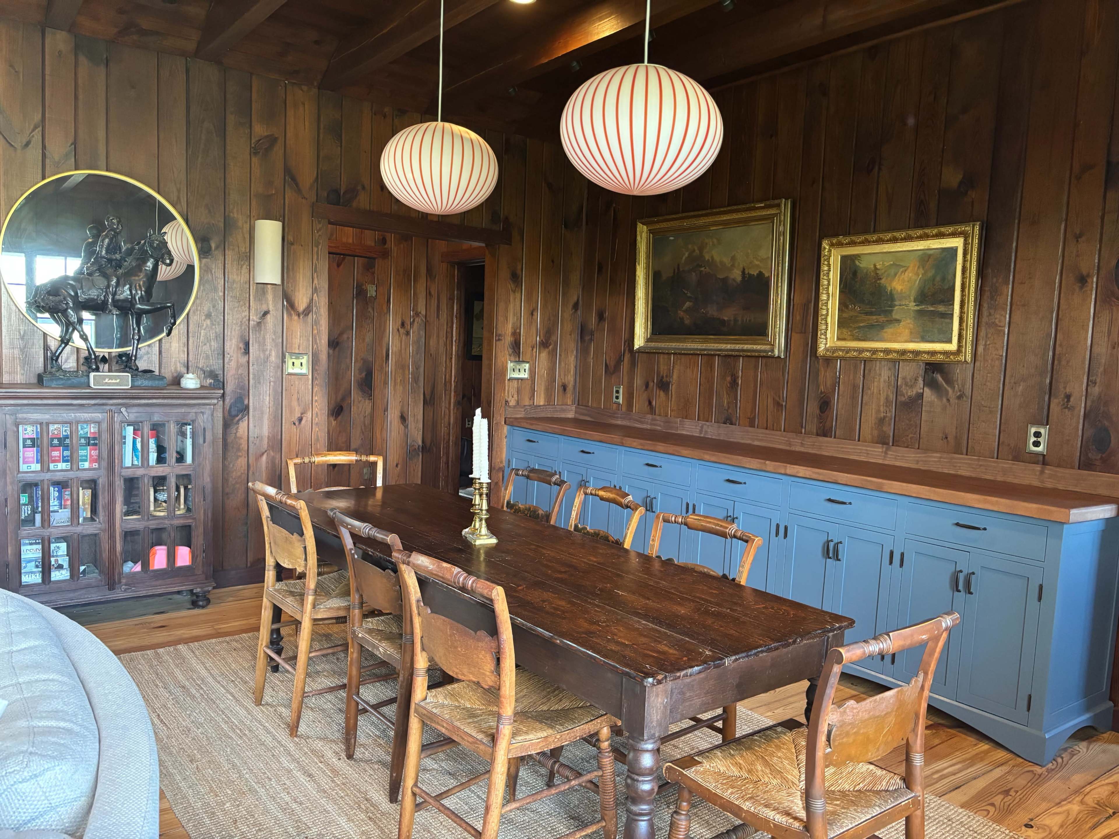 A dining area features a long wooden table surrounded by wooden chairs, with pendant lights hanging above and wood-paneled walls adorned with artwork.