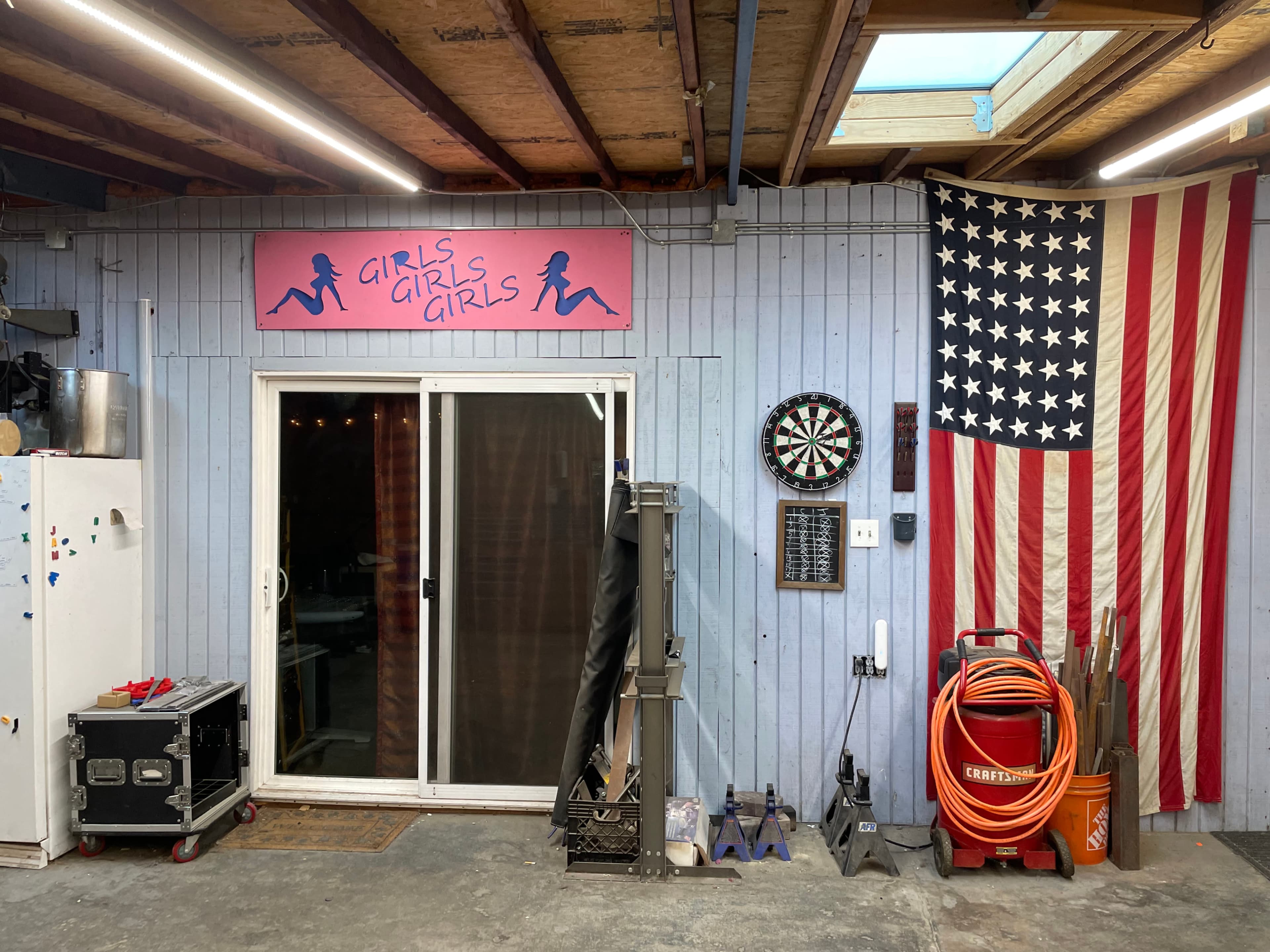 A wooden-paneled garage or indoor space with a sliding glass door, a dartboard, an American flag, and various tools and equipment arranged nearby.
