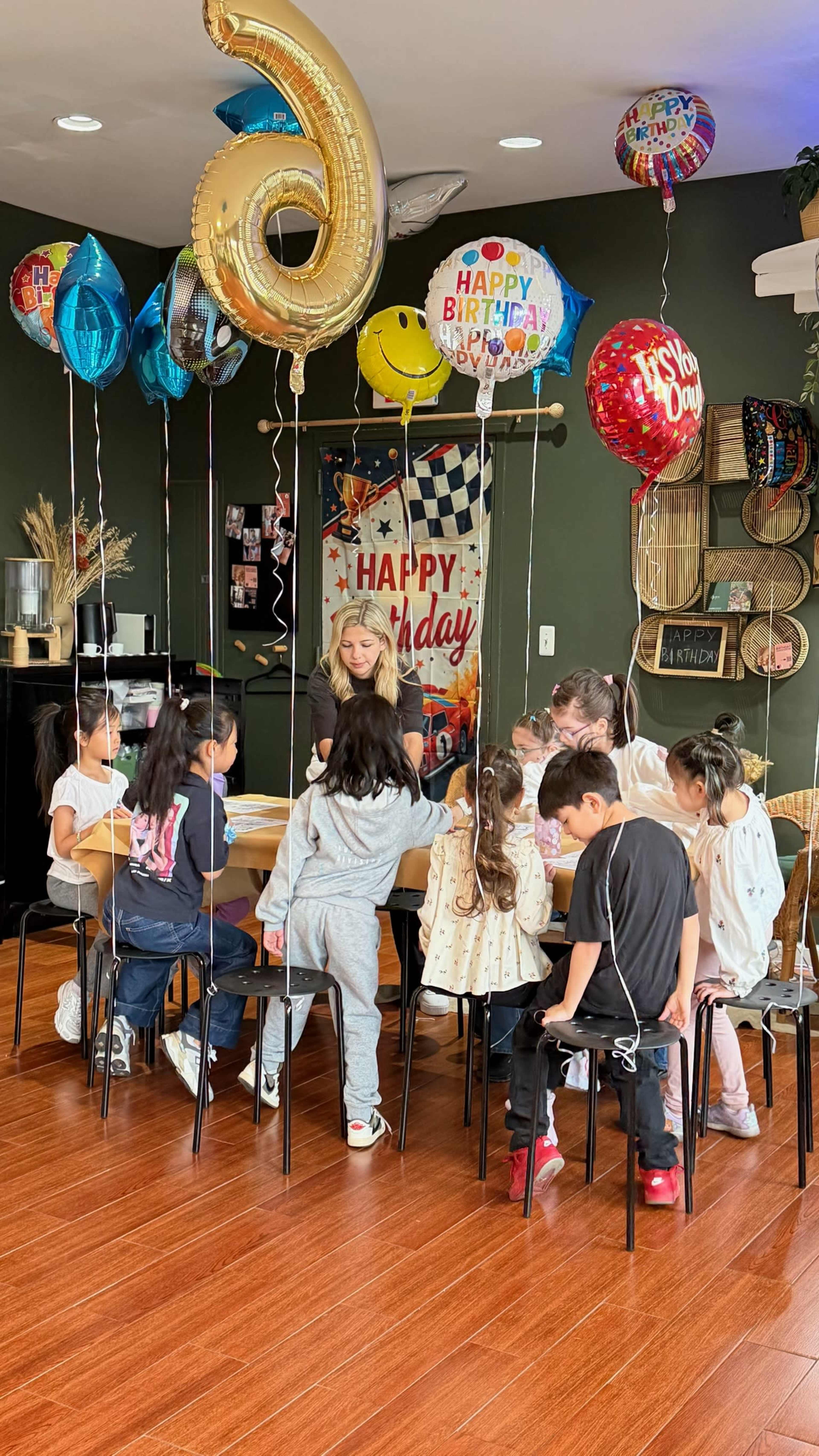 A group of children sits around a table decorated with birthday balloons, while an adult engages with them in a celebration setting.