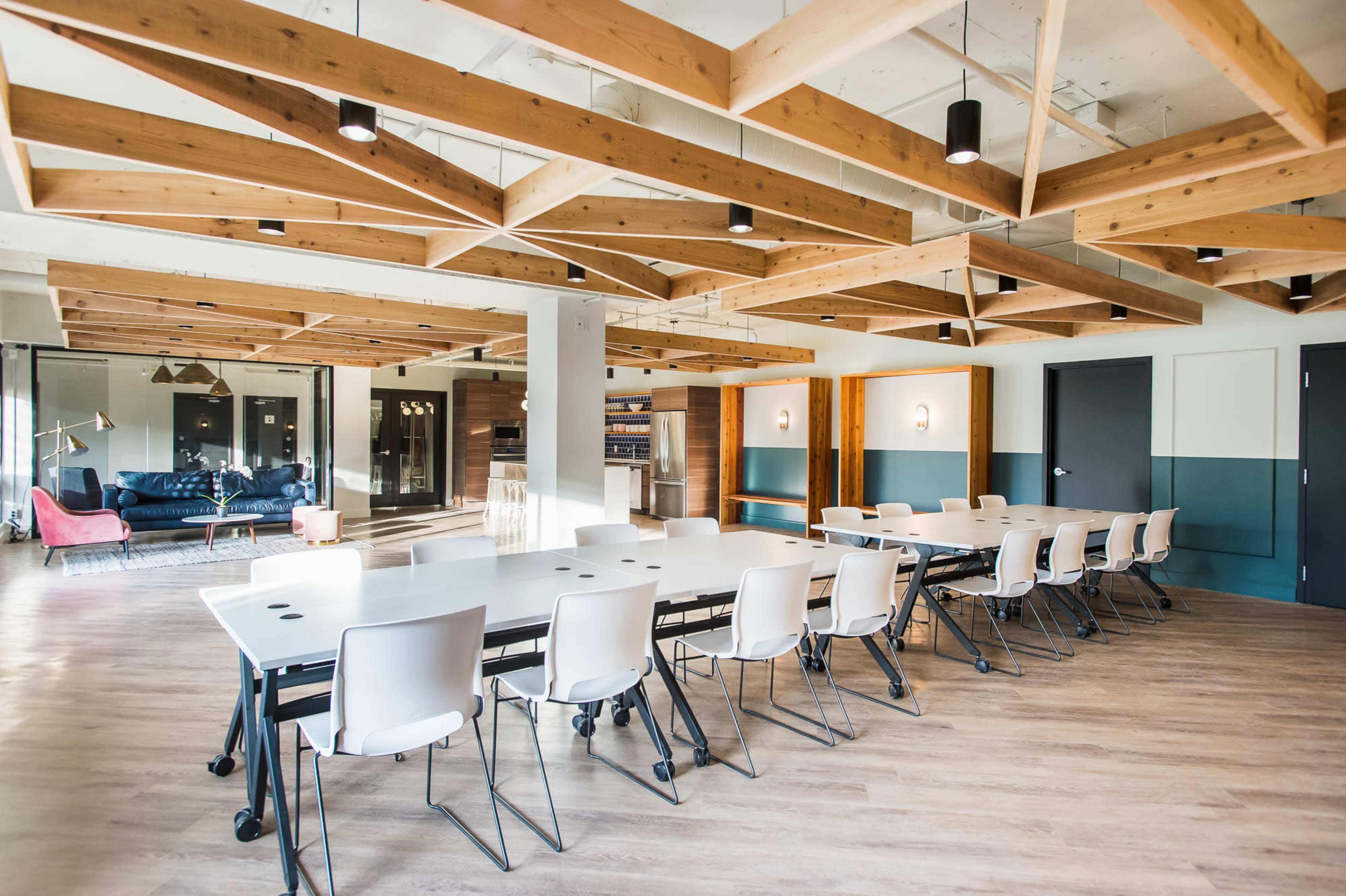 A modern conference room features a long table surrounded by rolling chairs, with wooden beam ceilings and large windows allowing natural light.