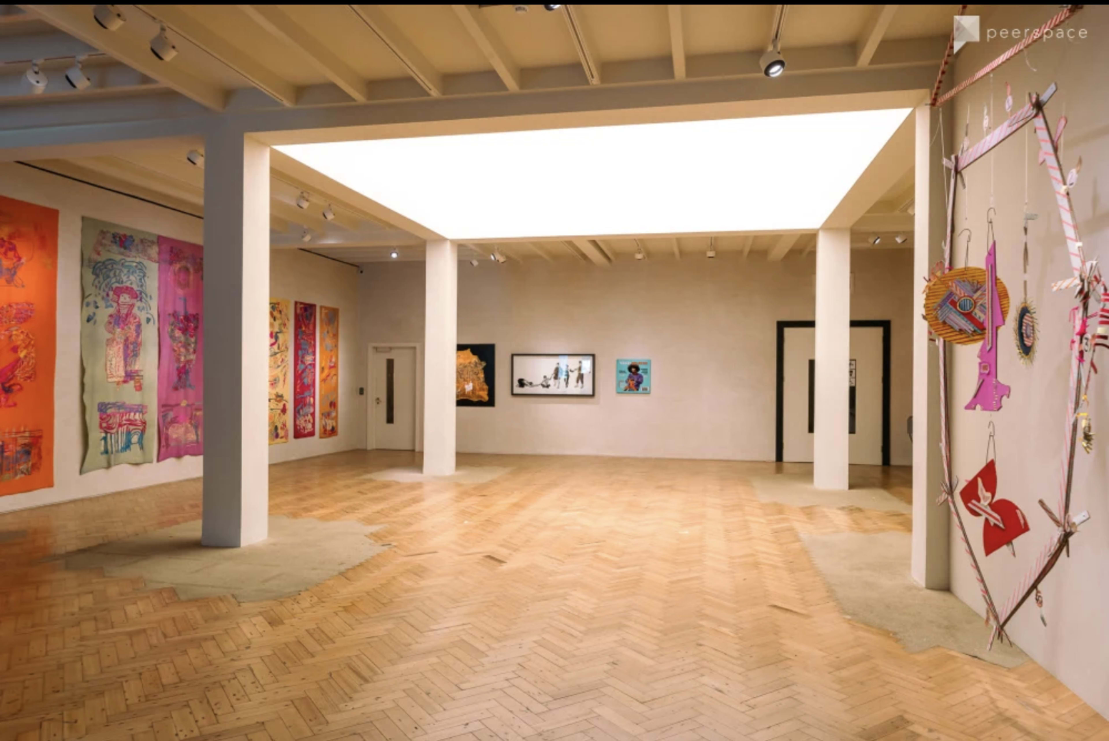 The interior of an art gallery features colorful wall hangings on two sides, with a central open space highlighting framed artwork and a skylight above.