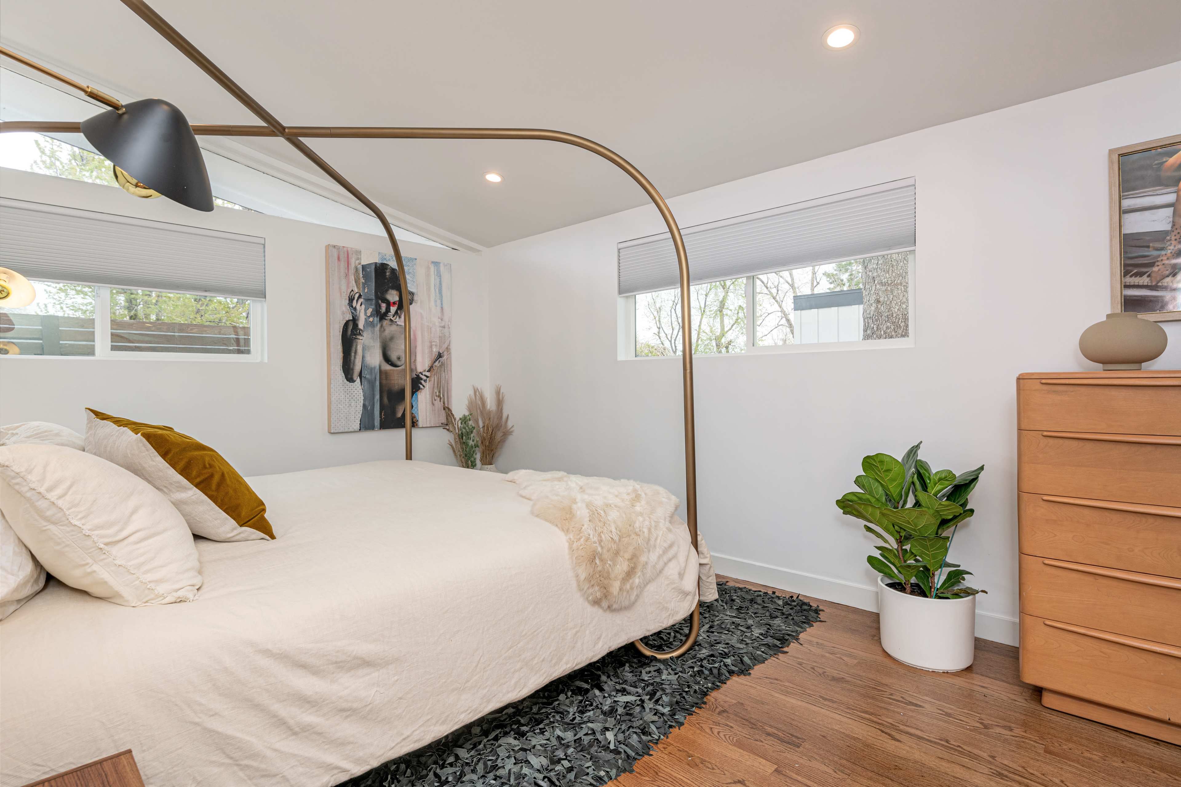 A modern bedroom with a brass bed frame, a neutral-colored bedspread, a potted plant, and a wooden dresser against the wall.