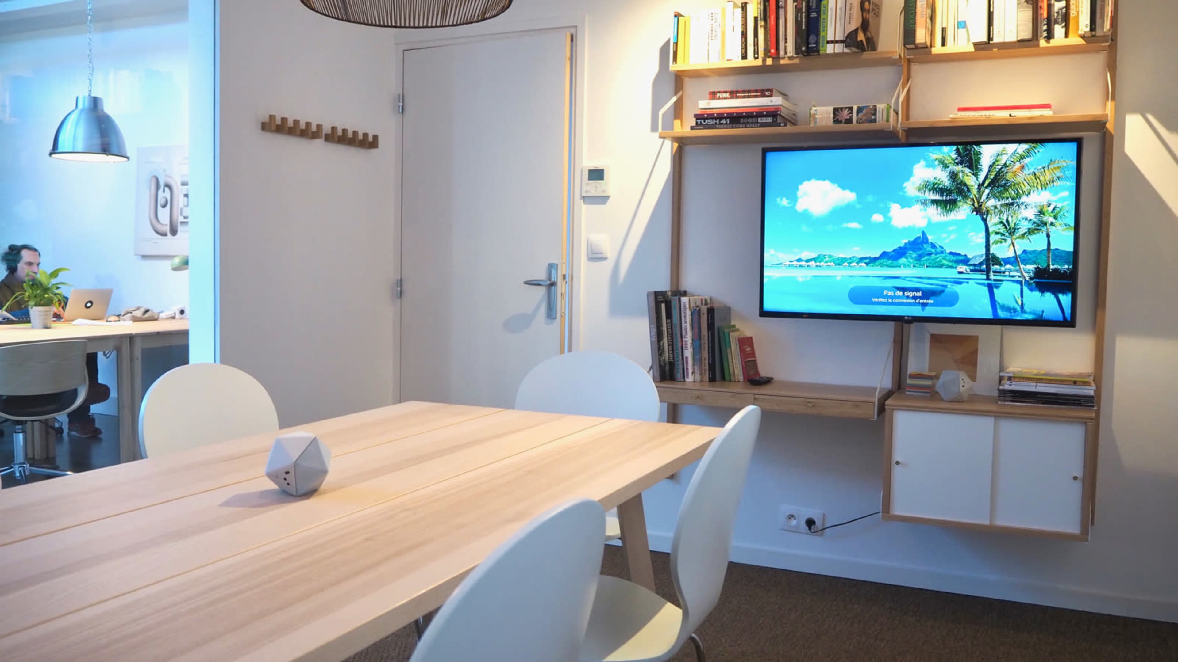 A well-lit modern living space features a wooden table, a wall-mounted TV displaying a beach scene, and a bookshelf filled with books and decorative items.