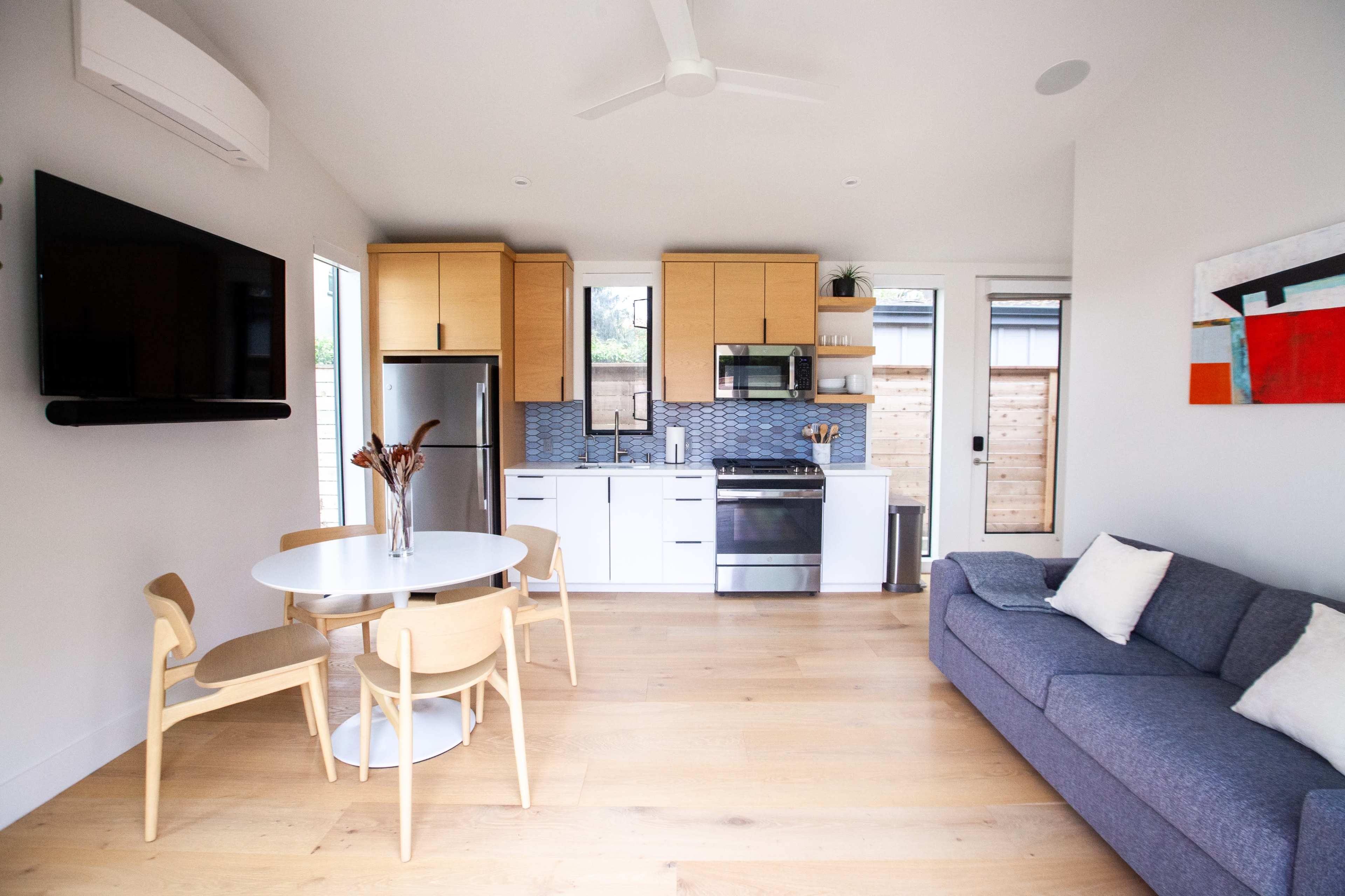 The image shows a modern, open-concept kitchen and living area featuring a dining table, chairs, a sofa, and kitchen appliances.