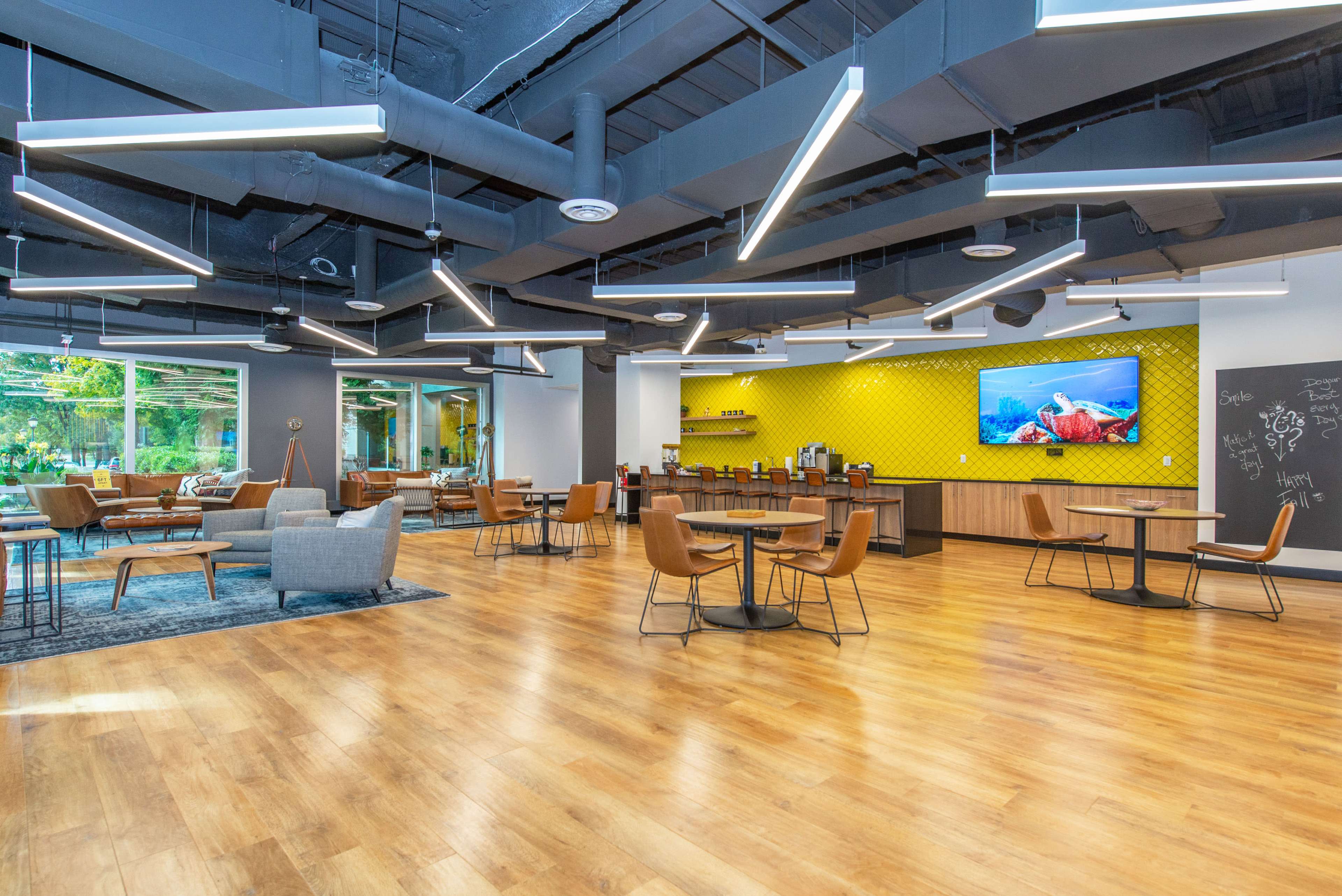The image shows a modern lounge area with wooden floors, seating arrangements, a coffee station, and large windows allowing natural light to enter.