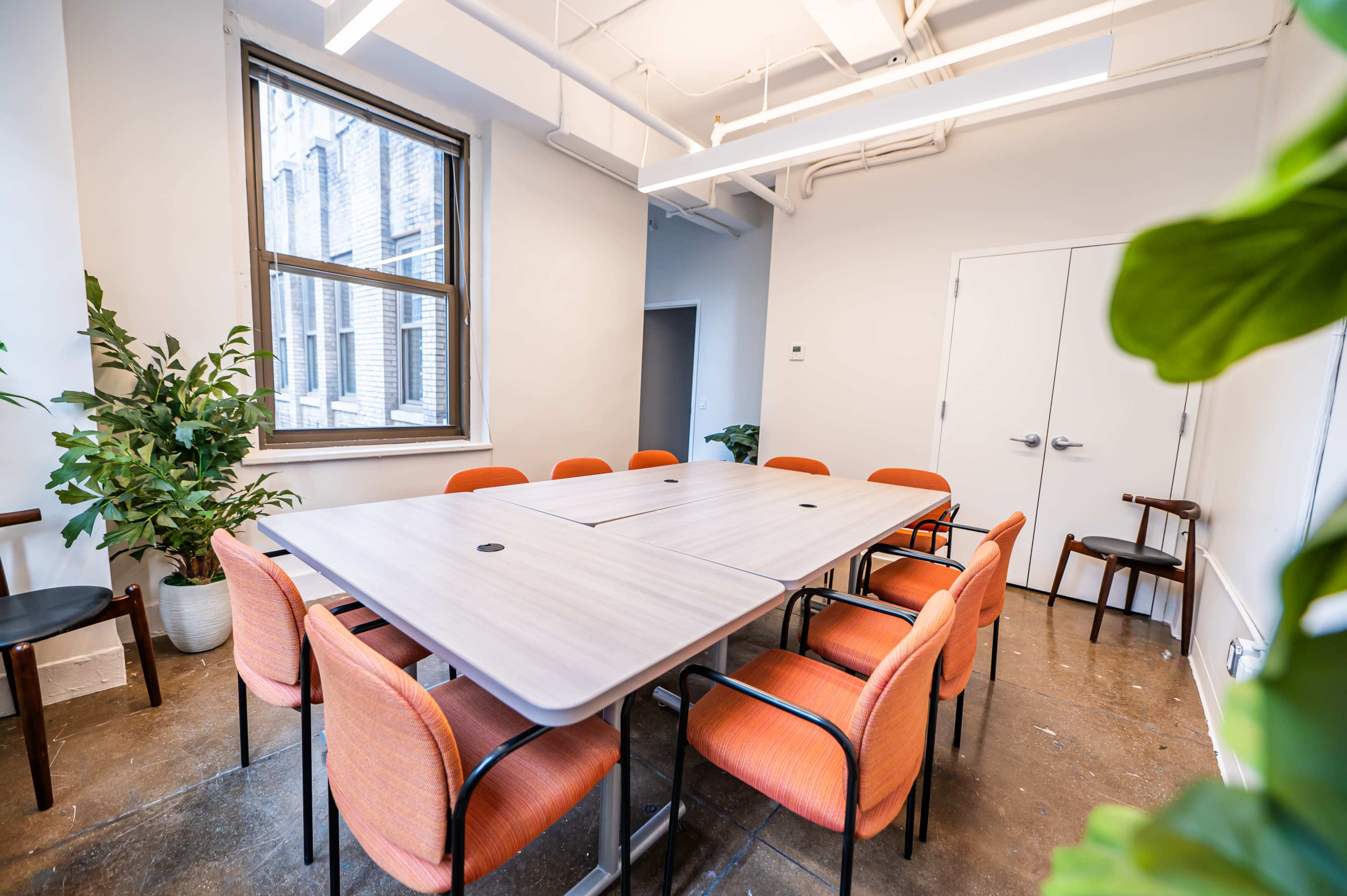 A conference room features a large rectangular table surrounded by orange chairs, with natural light coming through a window and potted plants in the corners.