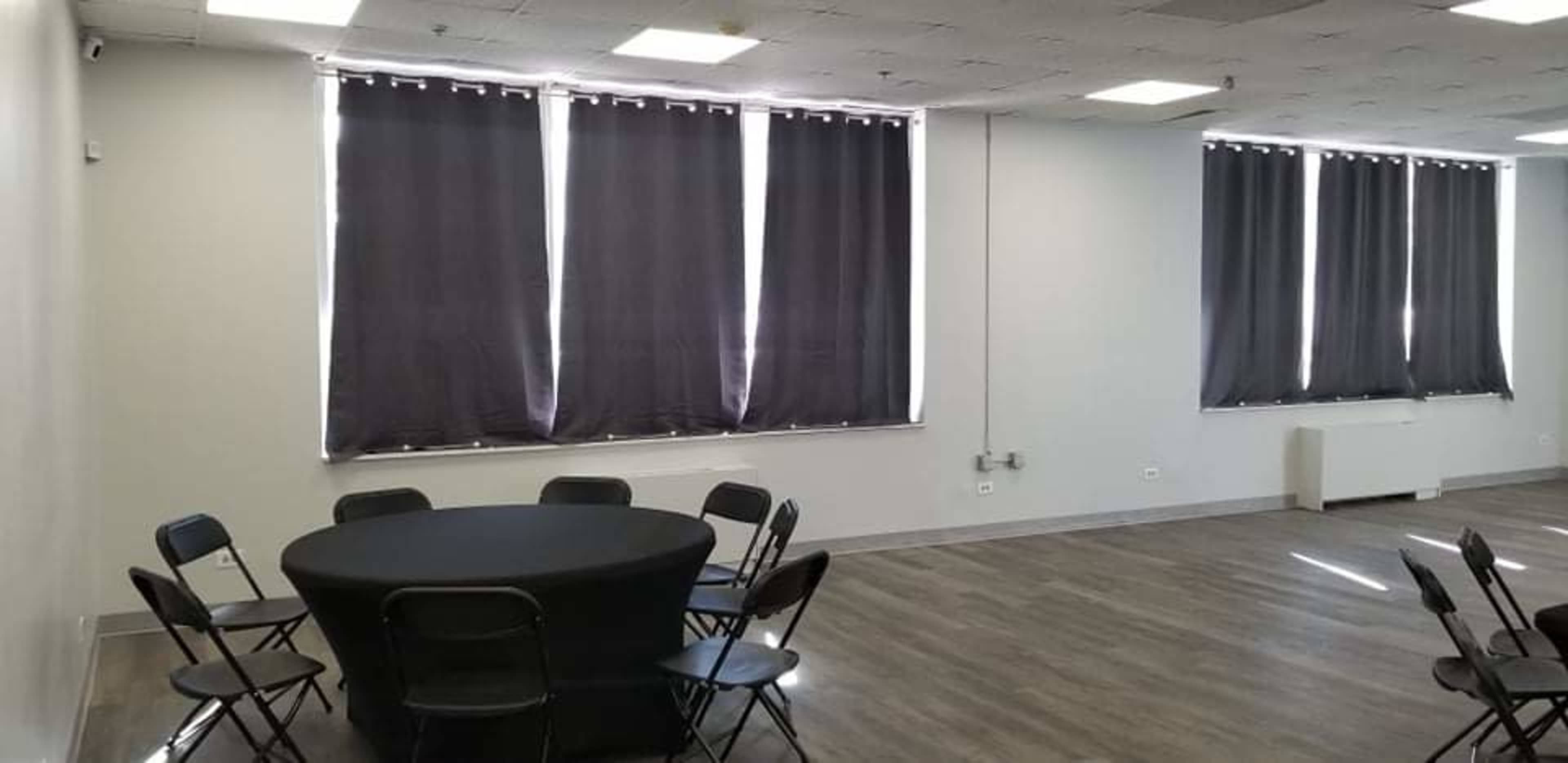 A simple meeting room features a round table surrounded by eight black chairs, with large windows covered by dark curtains.