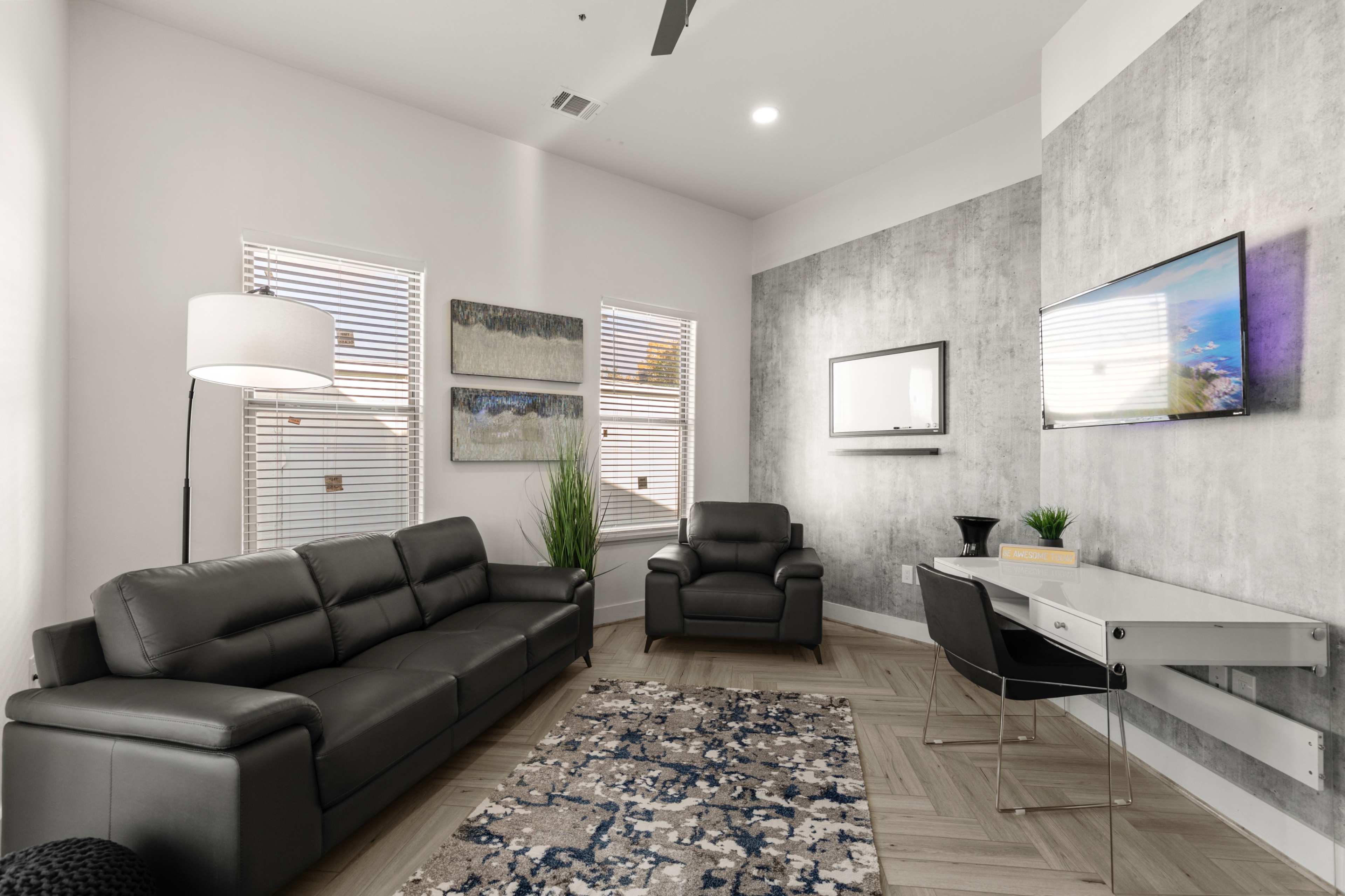 A modern living room features a gray sofa, an armchair, a desk, and a wall-mounted television, complemented by patterned rugs and potted plants.