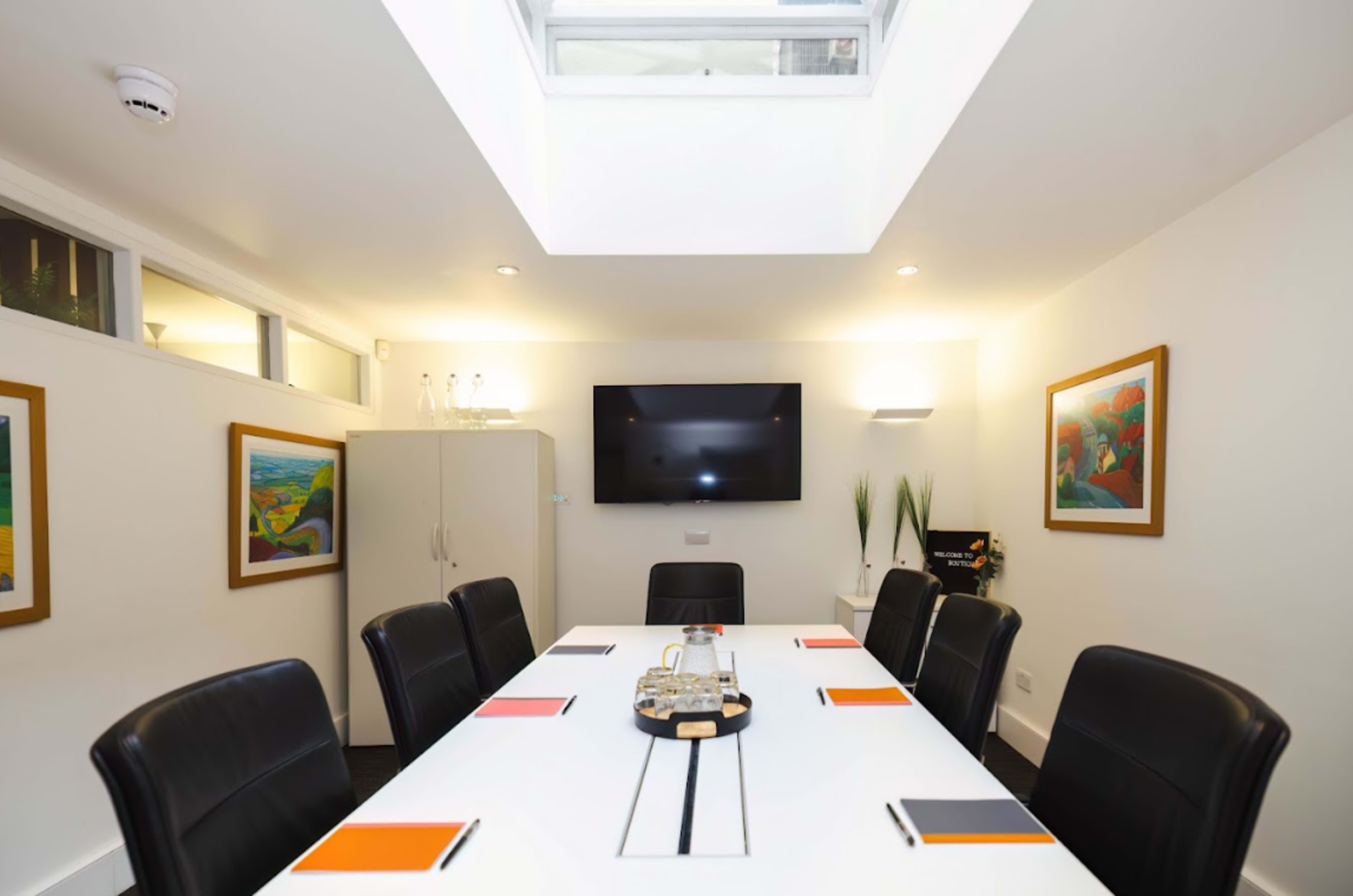 A modern conference room features a long white table surrounded by black leather chairs, with framed artwork on the walls and a television mounted on one wall.