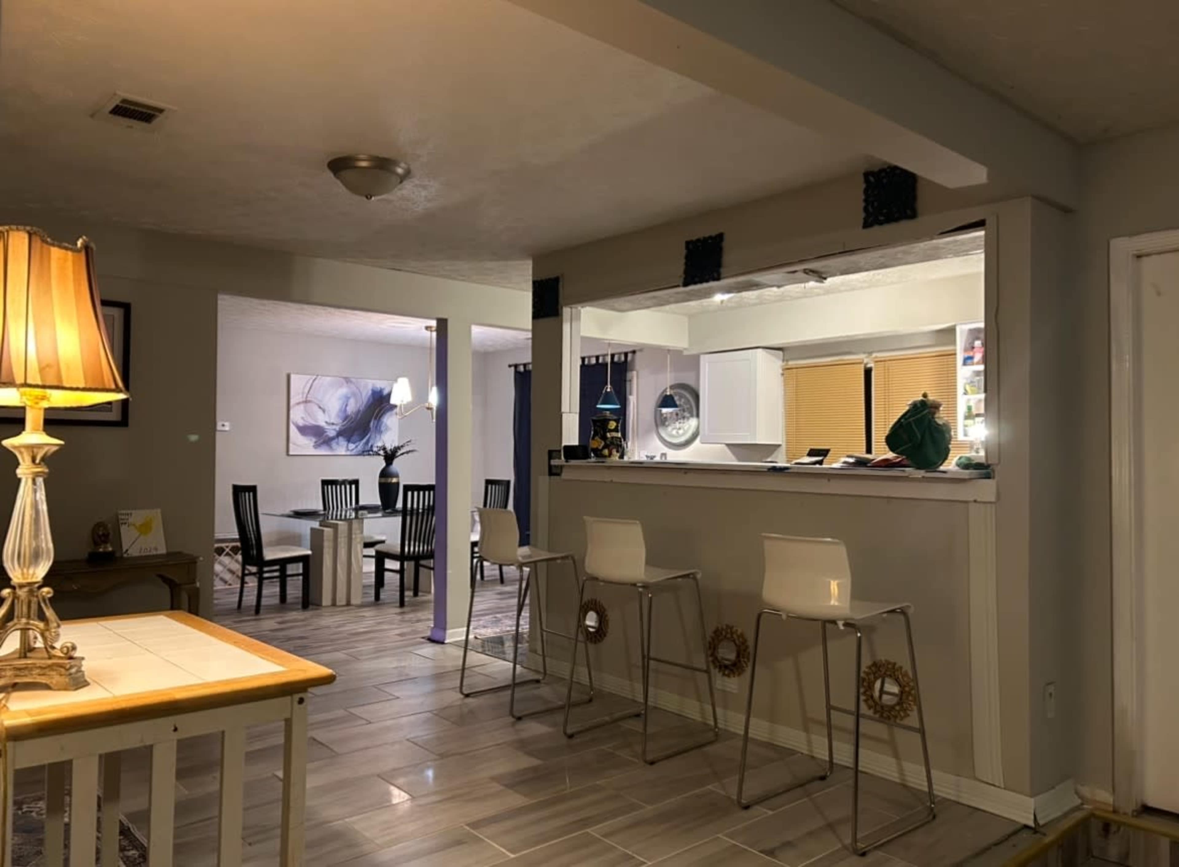 A living space with an open floor plan, featuring a kitchen bar with stools, a dining area with a table and chairs, and a soft light from a lamp.